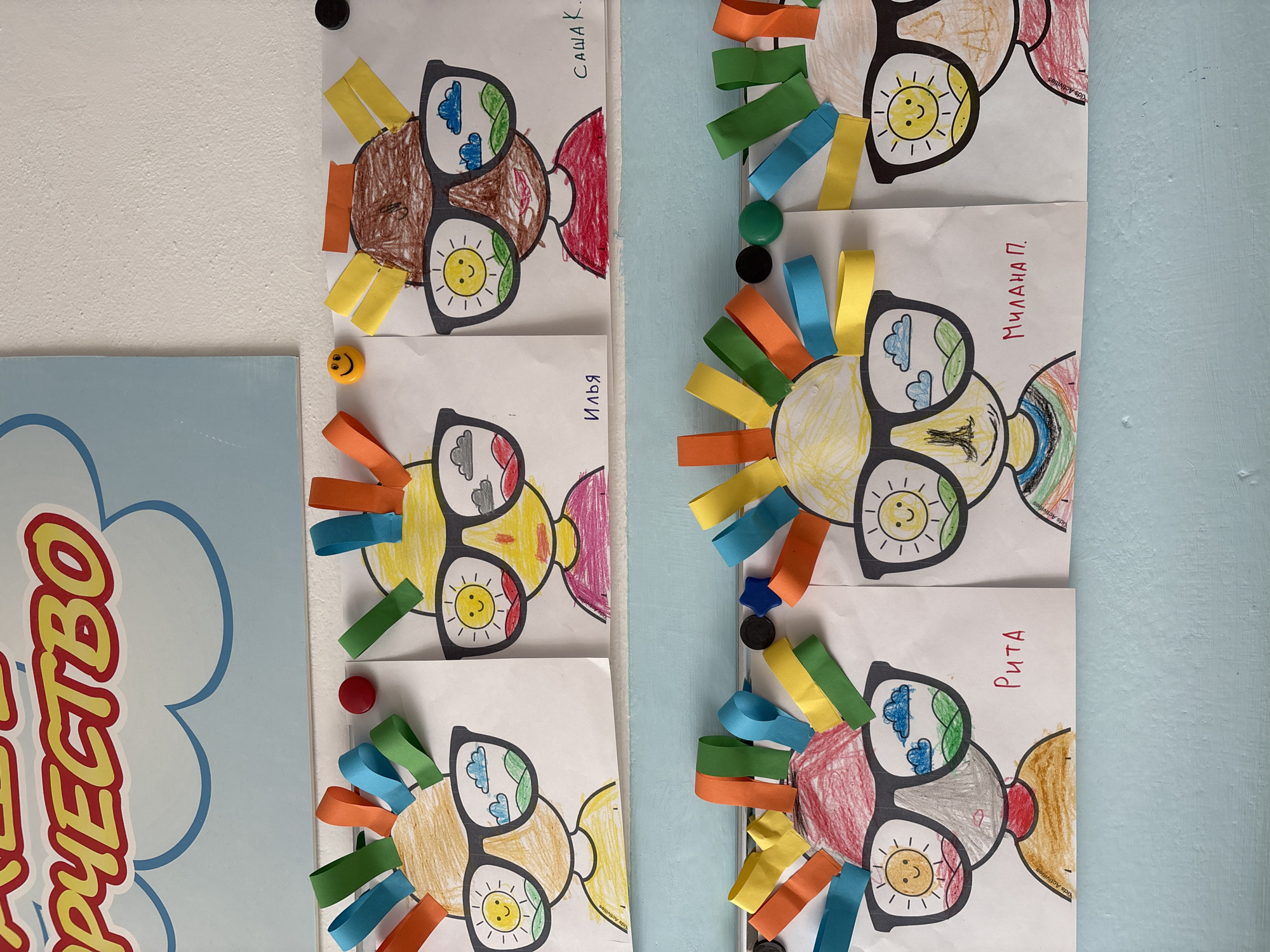 The image shows a wall decorated with children's artwork in what appears to be a classroom or community center. The artwork consists of portraits drawn on white paper, featuring children wearing oversized glasses. Inside the glasses, scenes of landscapes with suns and clouds are depicted. The heads of the children are decorated with colorful strips of paper resembling hair. Some of the artworks have names written below them in Cyrillic. Above the artwork, a portion of a sign with large yellow letters and a blue cloud design is visible. The wall itself is textured and white, with a blue shelf or ledge running horizontally across the lower part of the image. Magnets are used to attach the artwork to the wall. The overall impression is of a bright and cheerful display of children's creativity. While the image's exact location within Chaikovsky, Russia, cannot be determined, the artwork and the Russian names suggest a setting within Russia.