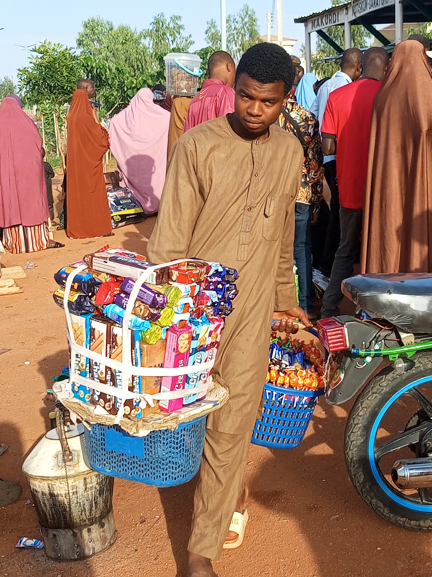 In an outdoor setting, a young man wearing a brown tunic stands in the foreground, looking towards the viewer. He is holding a large, blue basket filled with various packaged snacks, including cookies and candies, which are secured with white ties. Beside him, to his left, is a large, metal container with a lid and handle, resting on a circular base. To his right, a portion of a motorcycle is visible, featuring a black seat and a rear wheel with blue trim. In the background, several people are gathered, some wearing traditional Islamic clothing, including a woman in a pink hijab and another in a brown abaya. There is also a sign above a covered structure in the background that reads "MAKURDI KUTSKUN-UAKATORO". The ground is covered in reddish-brown dirt.