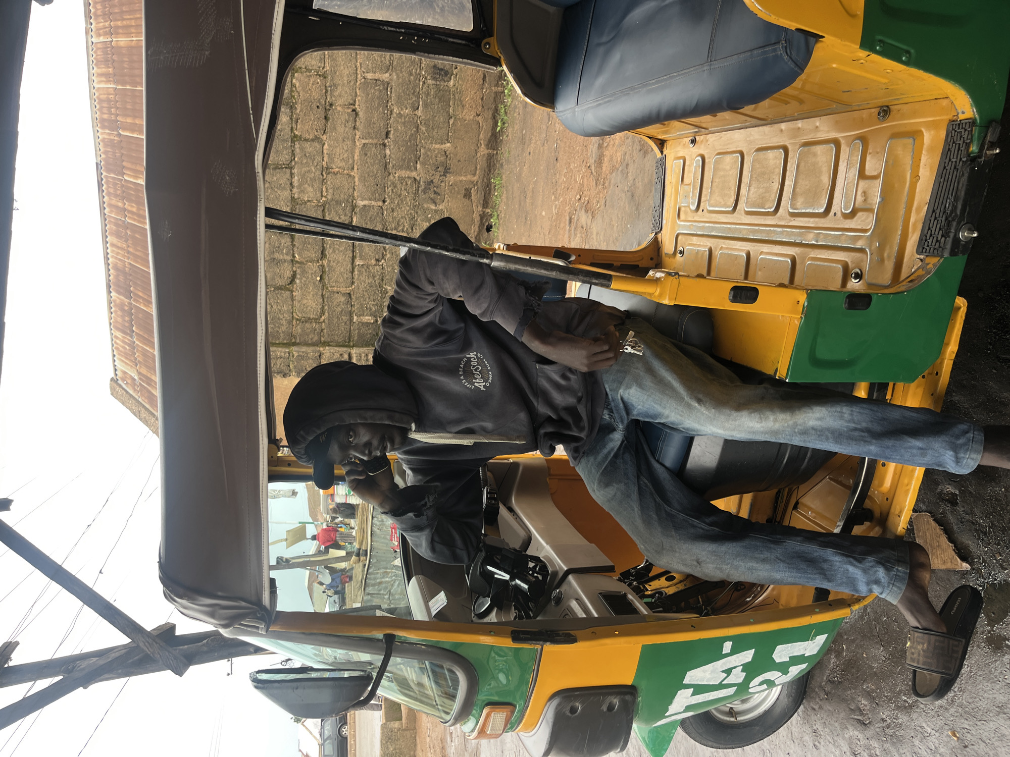 A man sits in the driver's seat of a yellow and green auto-rickshaw, holding a cell phone to his right ear with his right hand. He is wearing a dark hooded sweatshirt, blue jeans, and a black slide sandal on his left foot. The auto-rickshaw is parked on a dirt and pavement surface under an overhang. In the background, a brick wall is visible, along with other vehicles and people. The sky is overcast. The scene appears to be in Bukuru, Nigeria.