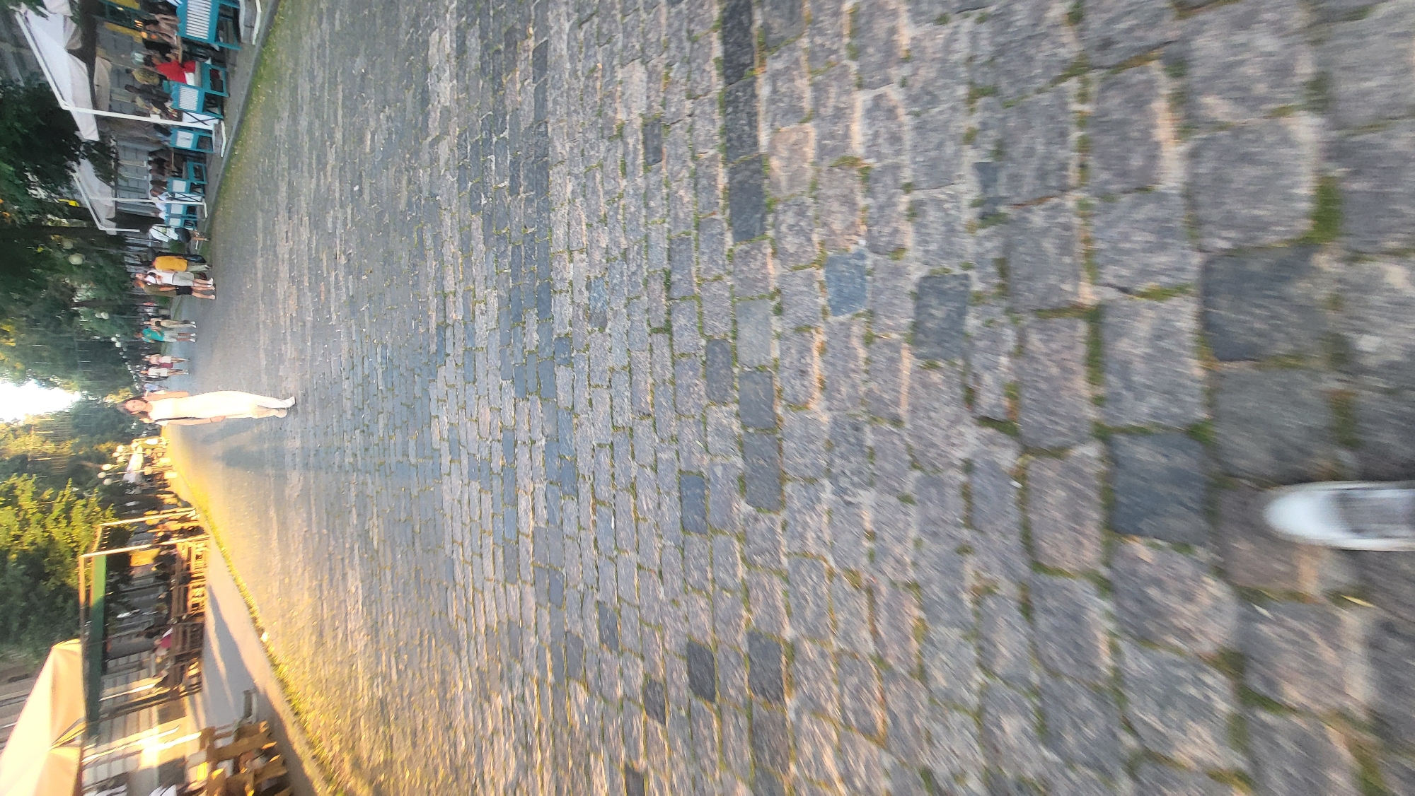 In this image, a woman in a white dress stands on a cobblestone path, her body angled towards the viewer. The path is lined with trees and a building with outdoor seating on the left side. Several other people are visible in the background, some walking and others seated at tables. The lighting suggests it is either early morning or late afternoon, with long shadows cast on the cobblestones. The overall atmosphere appears to be relaxed and leisurely, perhaps a park or a pedestrian walkway in a European-style town. The cobblestones are dark gray and appear well-worn, with small patches of grass growing between them. The building on the left has blue tables and chairs, and white umbrellas are set up for shade. The trees are lush and green, indicating a warm season. There is no visible text in the image.