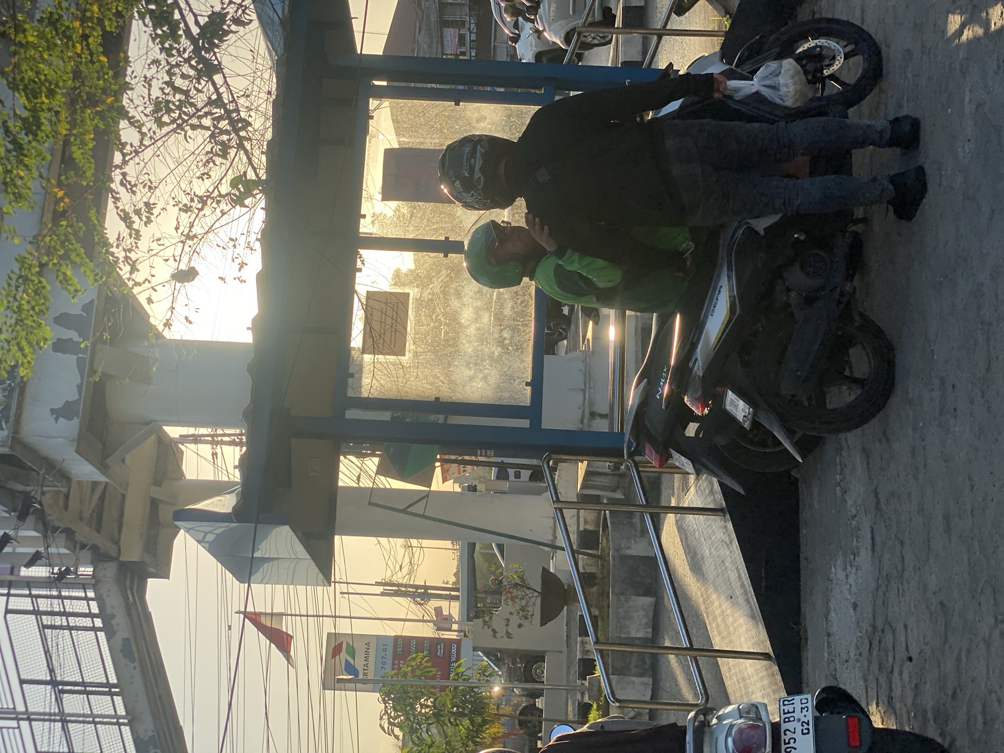 The image depicts a street scene in Banjarbaru, Indonesia, likely during sunset due to the warm, low-angled sunlight and long shadows. In the foreground, two individuals are standing next to motorcycles. One person wears a green jacket and helmet, while the other wears a black jacket and helmet, holding a plastic bag filled with items. They appear to be engaged in conversation. Several motorcycles are parked in the scene, including one with a license plate "952 BER G2.3G". Behind them, a bus stop shelter with blue framing and glass panels is visible. Further back, a Pertamina gas station sign and an overpass structure with metal railings are present. The overall environment suggests a public area within the city, possibly near a transportation hub or commercial district. Trees with foliage are interspersed throughout the scene, and power lines are strung across the sky.