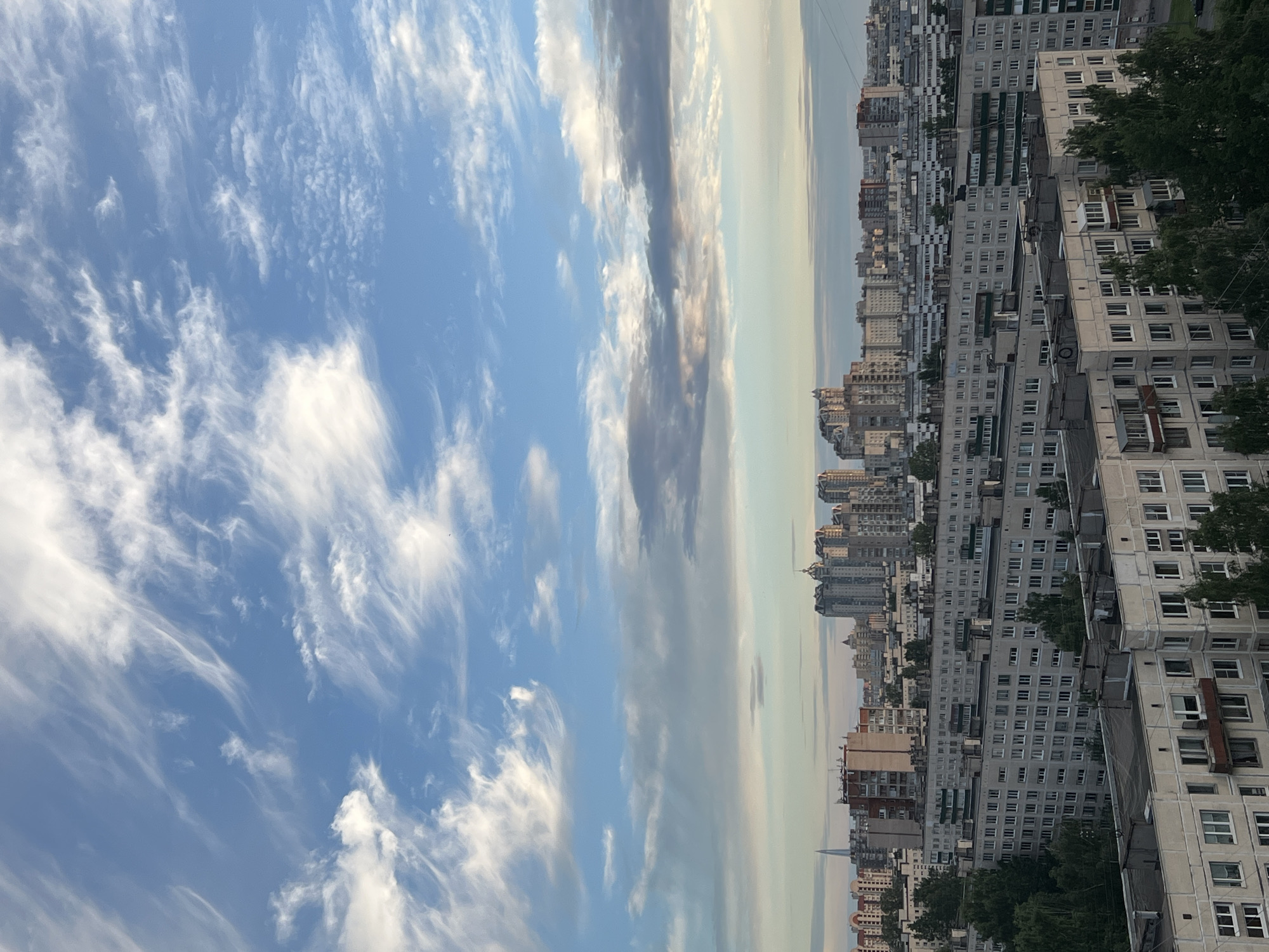 The image depicts a panoramic view of a cityscape in Sinyavino, Russia, with a striking contrast between a vast, dynamic sky and densely packed apartment buildings. The sky dominates the left side of the frame, filled with wispy, white cirrus clouds against a bright blue backdrop. These clouds are streaked and elongated, suggesting a breezy atmosphere.
On the right side of the image, a long line of multi-story apartment buildings stretches across the horizon. These buildings are predominantly grey and beige, with numerous windows, balconies, and some visible air conditioning units. The architecture suggests a typical Soviet-era housing complex. Interspersed among the buildings are patches of dark green trees, adding a touch of nature to the urban landscape. The buildings are tightly packed, creating a sense of density and uniformity.
The horizon line is slightly hazy, blending the sky and the distant cityscape. A faint, tall, spire-like structure is visible in the far distance, possibly a monument or a tall building. The lighting suggests it is either early morning or late afternoon, with the sun casting a soft glow and the sky still bright. There is no visible text in the image. The overall mood is one of urban sprawl under an expansive, cloudy sky.