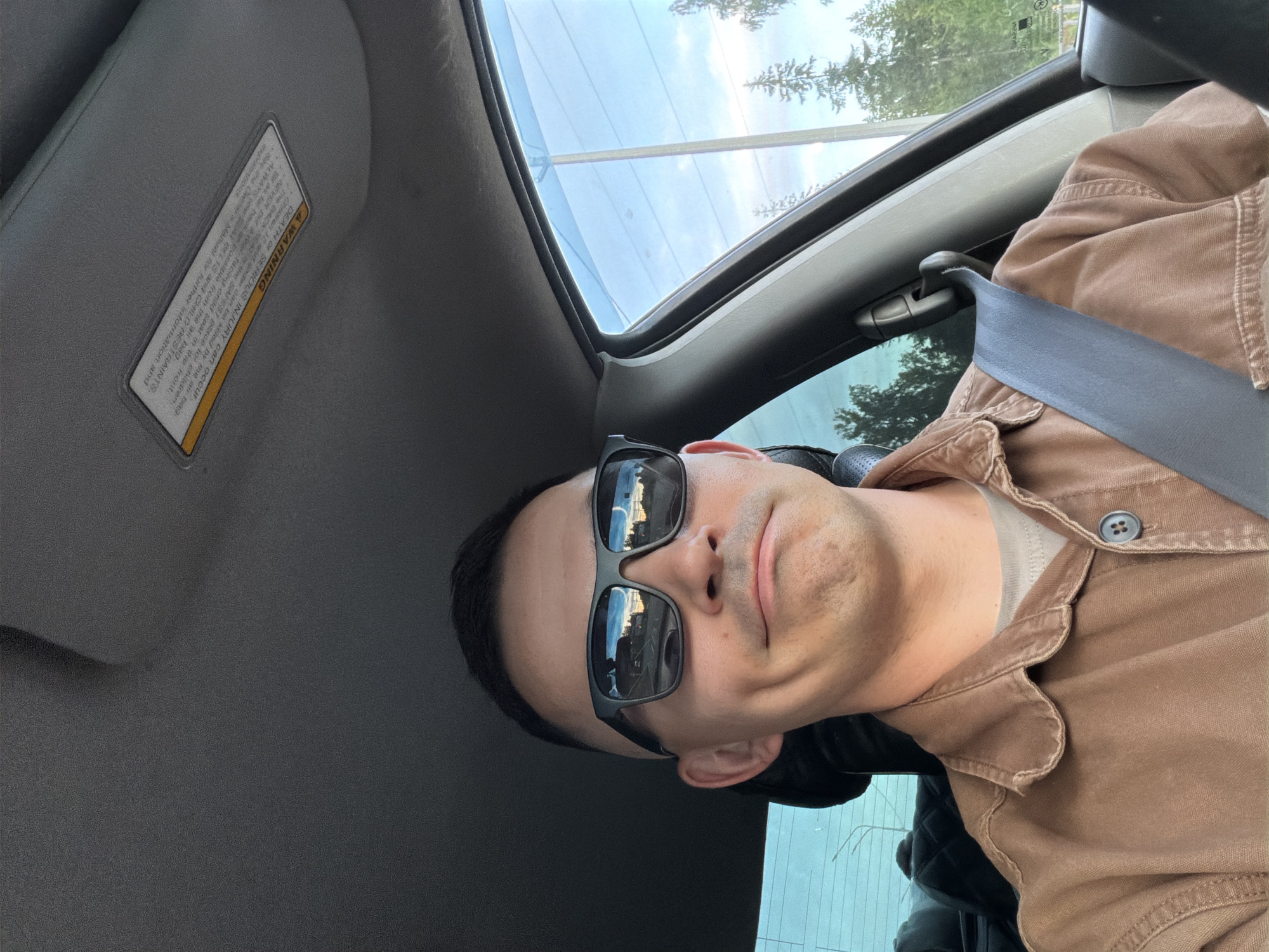 Here is a description of the image:
A man wearing sunglasses and a brown collared shirt sits in the driver's seat of a vehicle. He has his seatbelt on. The car has a sunroof. There is a warning sticker on the sun visor. The surrounding environment is visible through the car windows and sunroof, showing trees and the sky. The picture may have been taken while the car was stationary. There is no indication of Severnyy, Russia in the image.