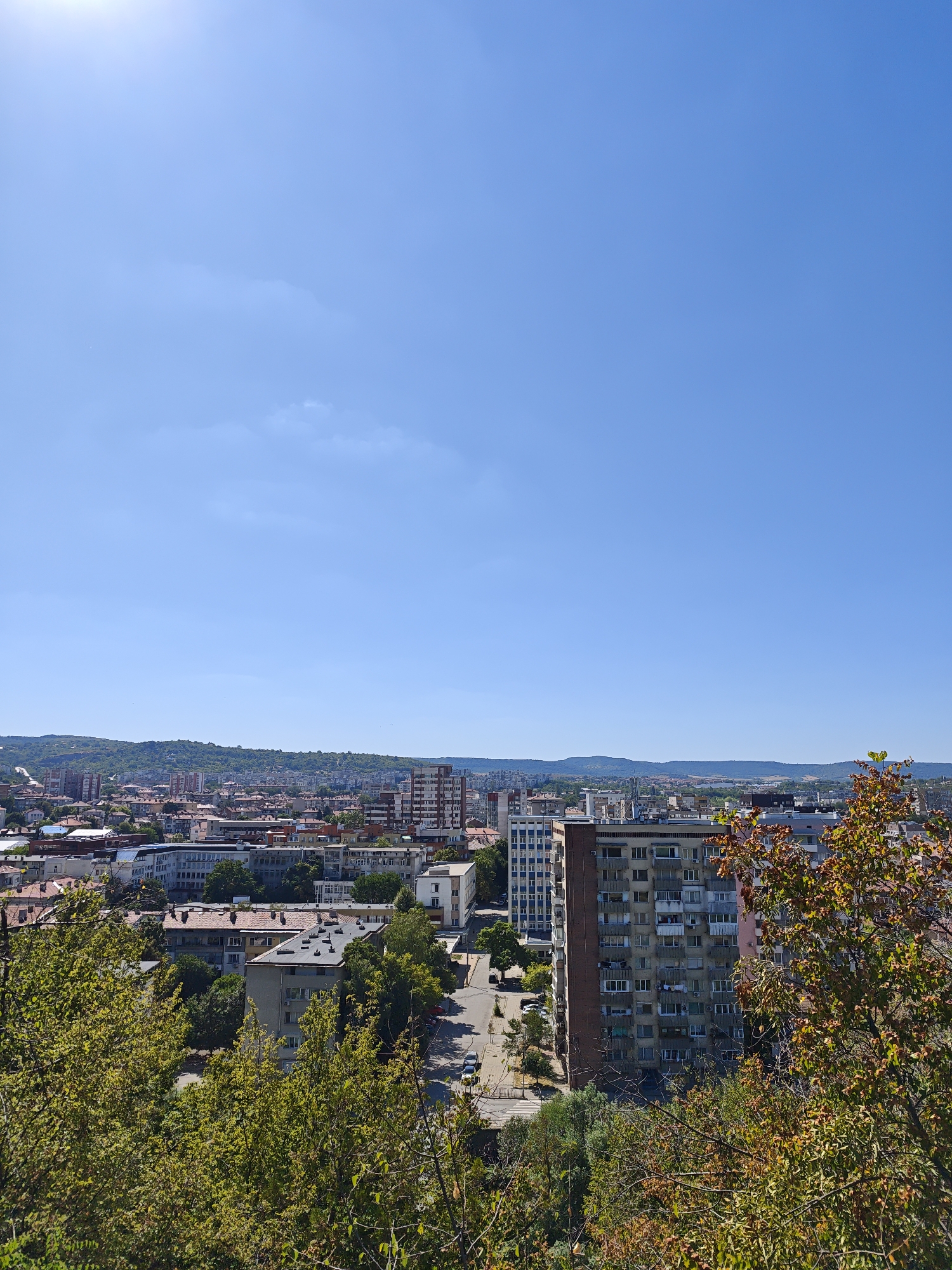 This is a wide-angle, eye-level, outdoor, daytime photograph taken from an elevated position overlooking the city of Lovech, Bulgaria. The sky is clear and blue, with a few wispy clouds. The sun is visible in the upper left corner.
In the foreground, lush green trees with some autumnal colored leaves partially obscure the view of the city. The middle ground is dominated by a dense urban landscape, featuring a mix of residential buildings, some with red tiled roofs and others with modern facades, as well as taller apartment blocks. A street runs through the city, with a few parked cars visible. The background shows rolling hills and more distant urban sprawl, with a hazy, distant mountain range on the horizon. The architecture suggests a Soviet-era influence in some of the larger apartment buildings. There are no visible people in the image.