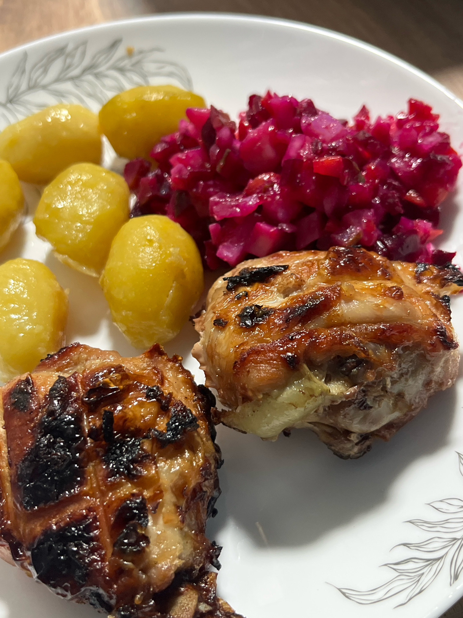 A close-up, overhead shot captures a white plate of food on a wooden table. The plate is decorated with a silver floral pattern. The meal consists of two pieces of grilled chicken, a pile of small, whole boiled potatoes, and a vibrant red salad. The chicken pieces are golden brown with visible char marks, suggesting they have been grilled or roasted. The potatoes are uniformly yellow and appear to be cooked until tender. The red salad, likely a beet salad, is finely diced and has a glistening texture. The lighting is natural and bright, highlighting the textures and colors of the food. The scene is a simple, close-up presentation of a cooked meal. There is no indication of the city of Nizhny Tagil, Russia, or any people in the image.
