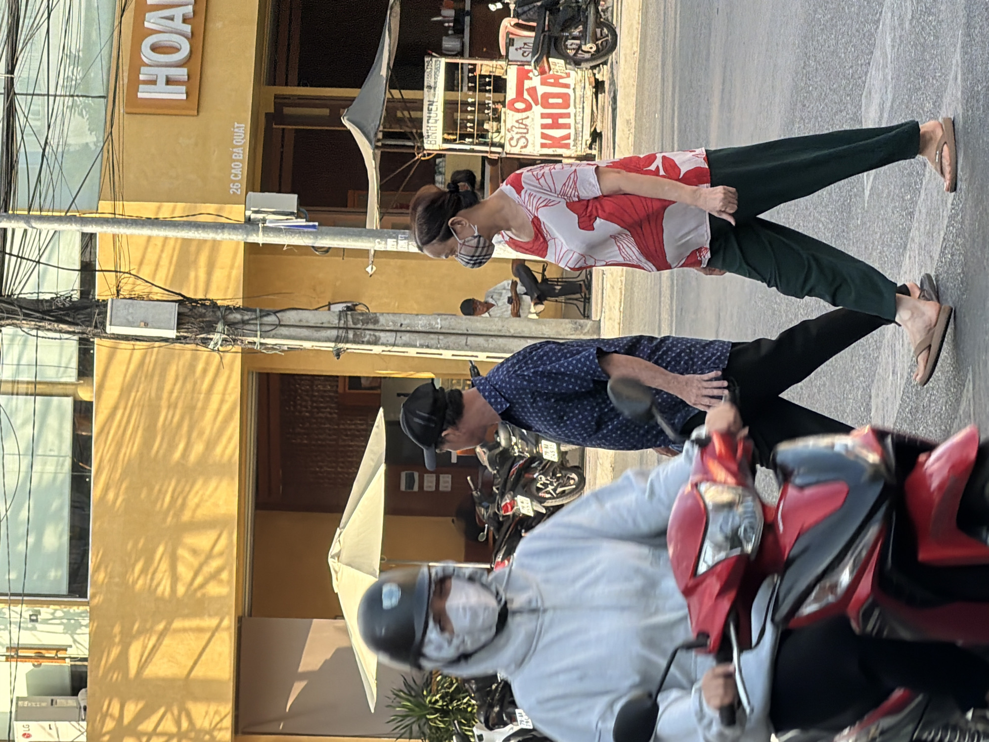 The image depicts a street scene in Nha Trang, Vietnam, under bright sunlight. In the foreground, a person on a red motorcycle is moving from left to right, partially obscuring the view. The rider is wearing a white hooded jacket, a black helmet, and a white face mask with a patterned overlay.
Behind the motorcycle, two pedestrians are walking across the street. The person on the left is a man wearing a dark blue patterned short-sleeved shirt and black pants. He is looking to his right. The person on the right is a woman wearing a white and red patterned top and dark green pants. She has dark hair pulled back and is wearing a checkered face mask. She is looking to her left.
In the background, a yellow building with the word "HOAN" visible on a sign is present. Below the sign, "26 CAO BA QUAT" is written. Several motorcycles are parked in front of the building. A street vendor's cart with various items and a sign that includes the word "SUA KHOA" is visible. The environment includes a street, sidewalk, utility poles with numerous wires, and trees casting shadows.