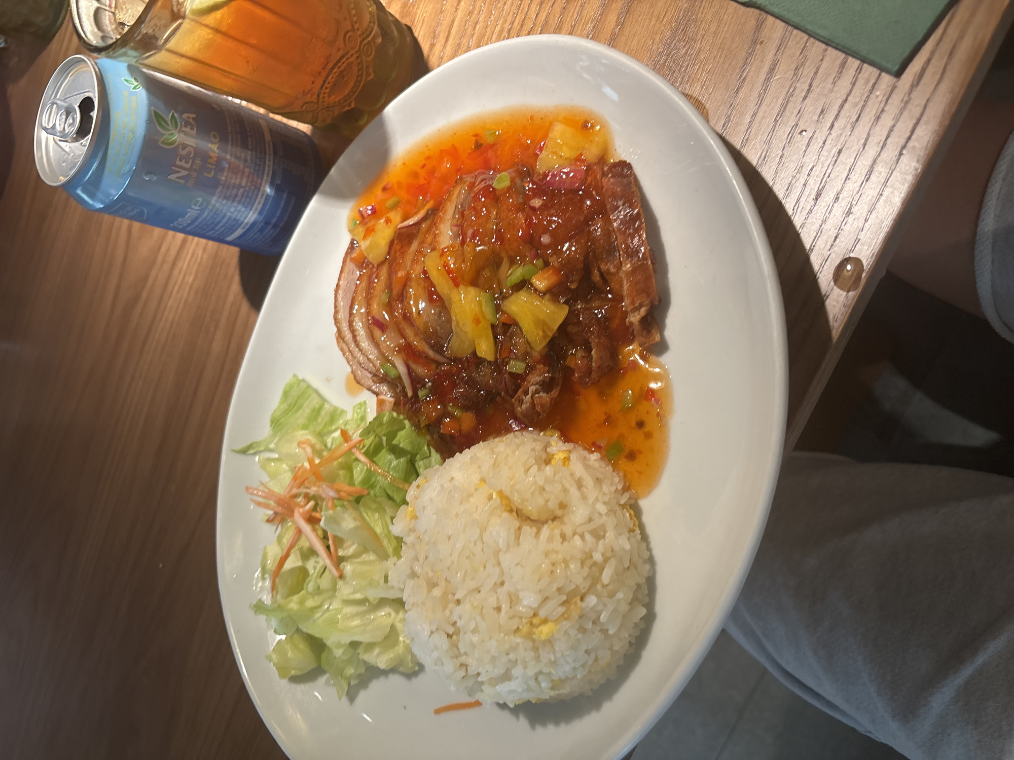 A white plate holds a meal of sliced duck breast covered in a glistening orange sauce with visible pieces of pineapple and red onion. To the left of the duck is a mound of white rice, and to the left of that, a small salad of shredded lettuce and julienned carrots. A can of Nestea Limão iced tea and a glass filled with iced tea and a lime wedge are positioned behind the plate on a wooden table. The lower portion of the image shows a person's legs, wearing light-colored trousers. The environment appears to be indoors, with the lighting suggesting daytime.