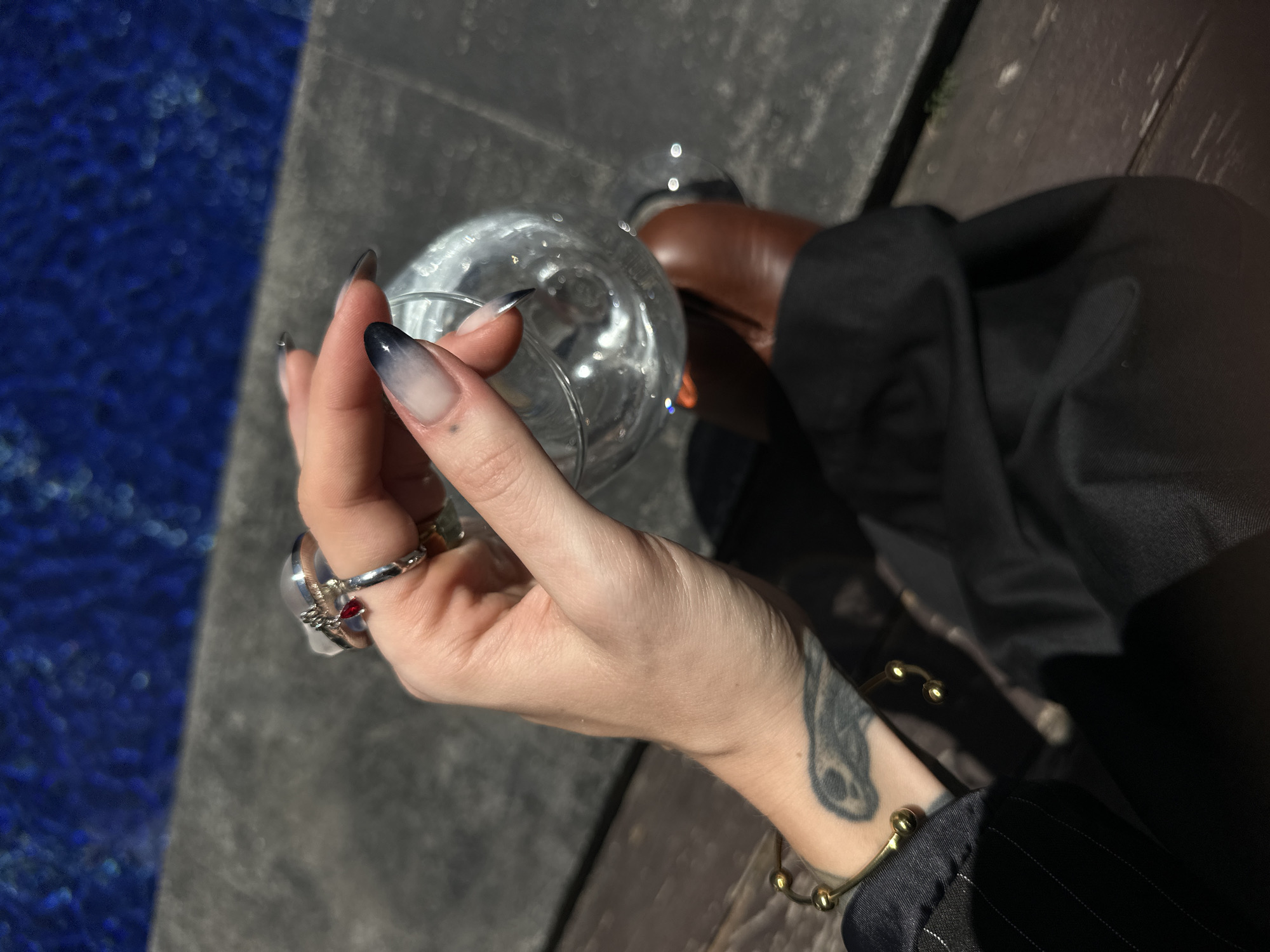 A person's hand with a dark-to-light gradient nail design is holding a clear glass filled with a clear liquid. The hand is adorned with several rings, including one with a red gem. A tattoo is visible on the person's wrist, and a gold bracelet is worn on the same wrist. The person is wearing a black jacket with pinstripes and black pants. Brown leather shoes are also visible. The background consists of a grey tiled surface and a body of blue water, suggesting an outdoor setting near a pool or ocean. The lighting indicates a sunny day. The scene is likely located in North Kuta, Indonesia, given the presence of tropical vegetation and architectural styles commonly found in the region.