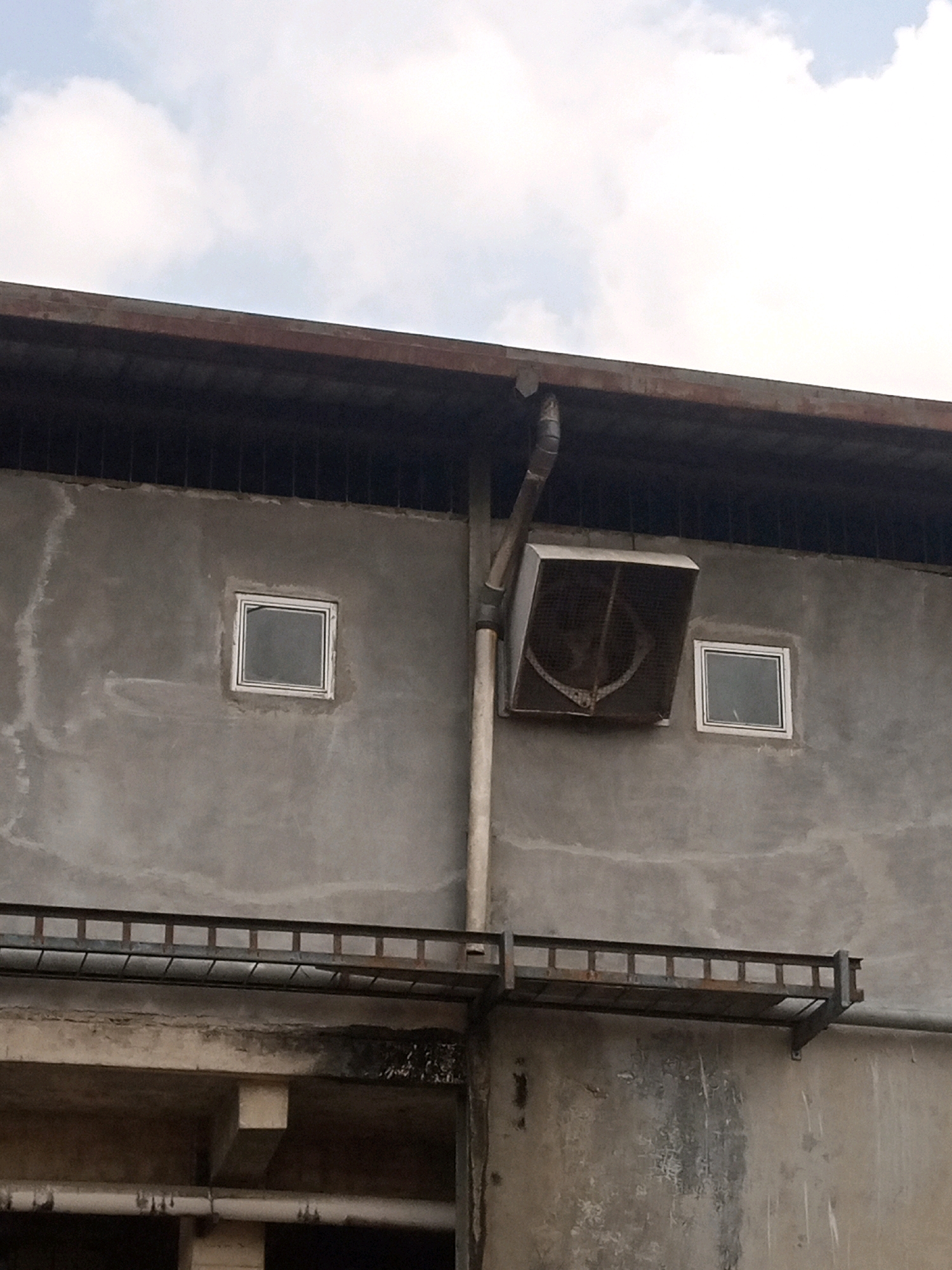 The image shows the exterior of a rough concrete building under a cloudy sky. The building has a dark, weathered overhang or roofline. There are two small, square windows with white frames on the concrete wall. A large, square ventilation unit with a metal grille is mounted on the wall between and slightly above the windows. A downspout runs vertically alongside the ventilation unit. A metal railing or walkway is attached to the wall below the windows. The lower portion of the image reveals structural elements of the building, including concrete beams and pipes. The scene appears to be in an industrial or utilitarian area. The location of Teluk Dalem Ilir, Indonesia is not visually represented in the image.