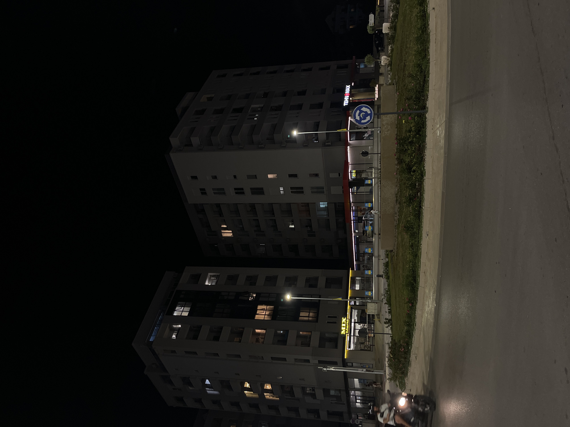 This is a nighttime photograph of a street in Budva, Montenegro, featuring two multi-story apartment buildings with illuminated windows. The building on the left has a sign that reads "MIX Markt." The building on the right has a sign that reads "TEHNO&MAX." In the foreground, a person is riding a scooter on a wet asphalt road. A circular road sign indicating a roundabout is visible on the right side of the image. The area appears to be a mixed-use development with commercial spaces at the ground level of the buildings.