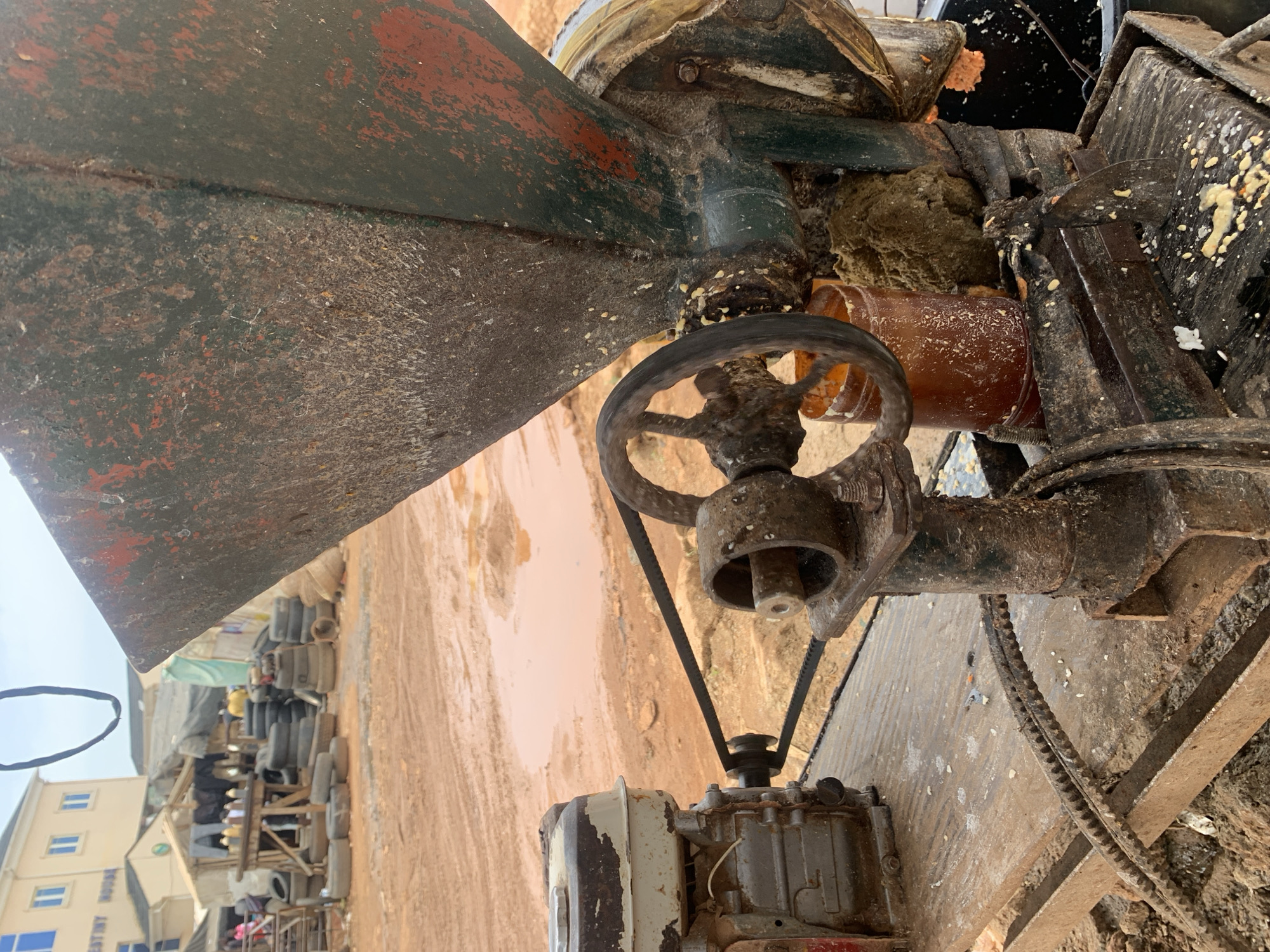 The image shows a machine in Dutsen Alhaji, Nigeria. The machine is a dark green and rusty piece of equipment with a large hopper at the top. It has a handwheel and a belt drive connected to a smaller motor near the bottom of the frame. The machine appears to be covered in residue. The background shows a dirt road and a building labeled "Destiny House". There are stacks of tires and other objects along the road, indicating a market area. There is evidence of people walking in the background near the market.