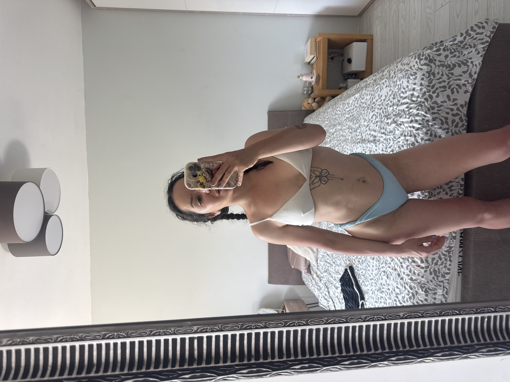 A woman is reflected in a mirror, taking a selfie with her phone. She is wearing a white bikini top and light blue bikini bottoms. She has dark hair styled in braids and visible tattoos on her arm and torso. She appears to be in a bedroom, with a bed covered in a patterned duvet visible in the background. A bedside table with some items on it, including a small white device and a pink cup, is also present. The room is lit by natural light, suggesting it is daytime. The overall mood appears casual and relaxed. The image is oriented vertically.