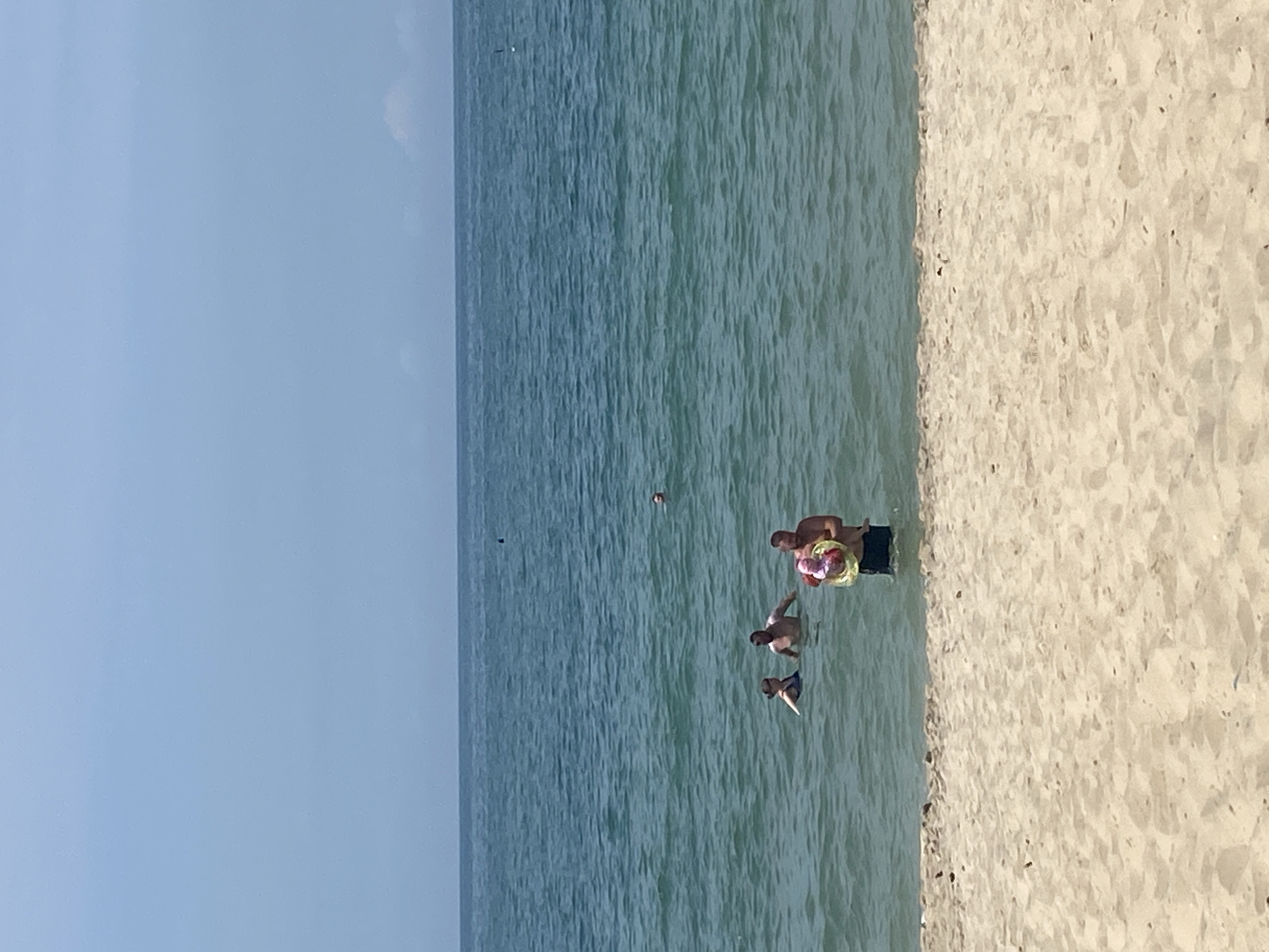 The image shows a group of people in the ocean on a sunny day. The water is a clear blue-green, and the sky is a pale blue. In the foreground, a sandy beach is visible. In the water, a man is holding a baby and a yellow inflatable ring. To his left, two other adults are swimming. Further out in the water, another person is visible. The scene appears to be a recreational area in Phu Thuy Ward, Vietnam.