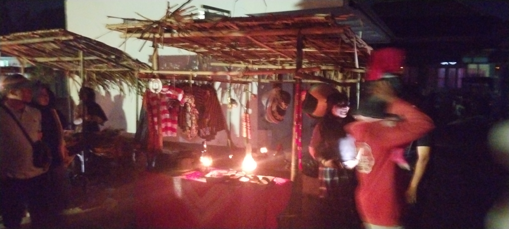 The image depicts a nighttime scene at a market stall in Keting, Indonesia. The stall features a thatched roof supported by wooden posts, with clothing items, possibly batik fabrics, hanging from a rack. Lit bulbs cast a warm glow on the displayed items and the surrounding area. Several individuals are present, some partially visible, suggesting a bustling atmosphere. One person in the foreground appears to be wearing a red garment, while others are in darker clothing. The background shows a dimly lit building with windows, indicating an urban or semi-urban setting. The overall impression is that of a vibrant local market at night.