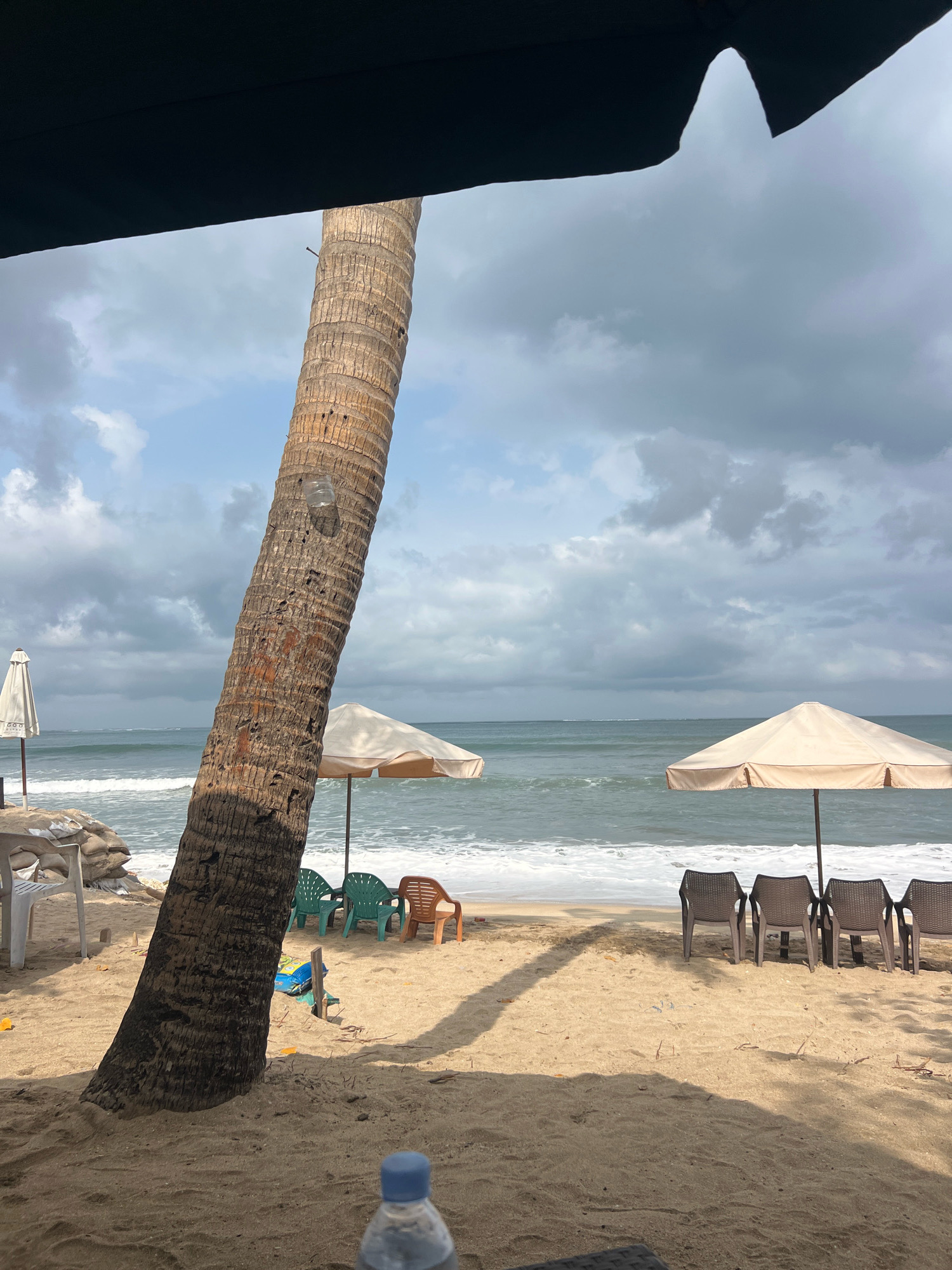 A tranquil beach scene unfolds in Kuta, Indonesia, under a sky filled with dramatic, cloudy formations. The foreground is dominated by the textured trunk of a leaning palm tree, its bark rough and weathered. To the right, two light-colored beach umbrellas stand, casting shadows on the sandy shore. Beneath them, a row of four dark plastic beach chairs are arranged facing the ocean.
The ocean itself is a dynamic expanse of blue-green water, with gentle waves creating a white foamy line as they approach the shore. The beach is a warm, golden sand, scattered with a few stray pieces of debris and a blue and yellow object near the base of the palm tree. On the left side of the image, a white beach umbrella stands in the distance, and several sandbags are piled nearby, suggesting some form of erosion control or protection. A white plastic chair is also visible. In the very foreground, out of focus, is a plastic water bottle with a blue cap. The overall atmosphere is one of a quiet, perhaps overcast, day at the beach.