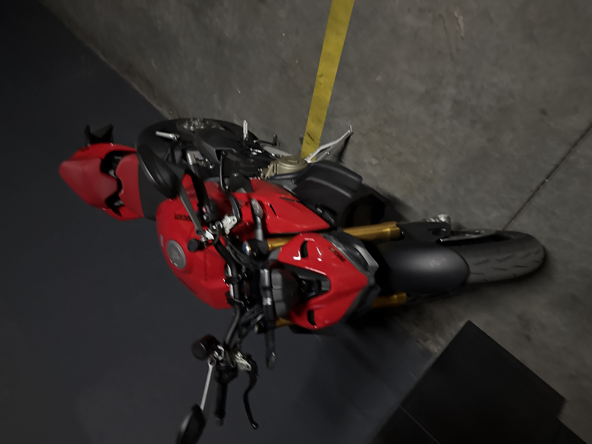 The image shows a red and black Ducati motorcycle lying on its side in what appears to be an indoor parking garage. The motorcycle is a sportbike with a sleek design, featuring a bright red fuel tank and fairings, contrasted with black accents and wheels. Gold-colored suspension components are visible. The motorcycle is positioned on a grey concrete floor, with a yellow line painted on the floor running vertically along the right side of the image. In the background, the garage walls are also grey concrete. The lighting suggests it is daytime, but the interior setting eliminates natural light as a primary source. No people are visible in the image, and there is no indication of a specific event or activity. The overall impression is a still life of a parked motorcycle in a utilitarian environment. Some faint text might be present on the motorcycle's fairing, but it is not clearly legible. The location context of Odesa, Ukraine, does not provide specific visual cues within the image itself.