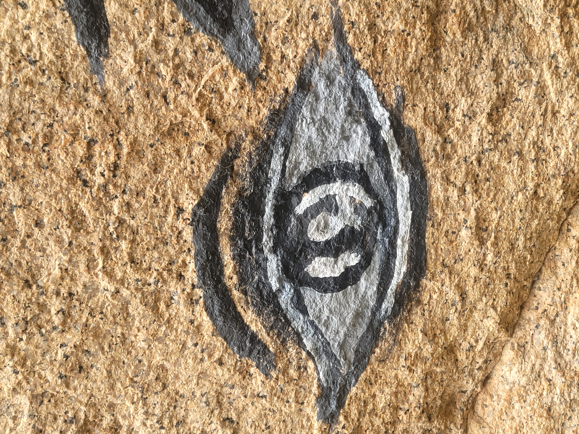 This image shows a close-up view of a painted eye on a textured, light brown, stone surface. The eye is rendered in shades of gray and white, with black outlines for the pupil and eyelids. The painting is stylized, with a simplified representation of the iris and pupil. The stone itself appears to be a coarse-grained granite or similar rock, with visible flecks of darker minerals. The texture of the stone is rough and uneven, providing a natural canvas for the artwork. The lighting suggests an outdoor environment, possibly during daylight. The specific location in Barakin Akawo, Nigeria, is not directly discernible from the image content itself, but the artwork is situated on a natural stone formation within that area.