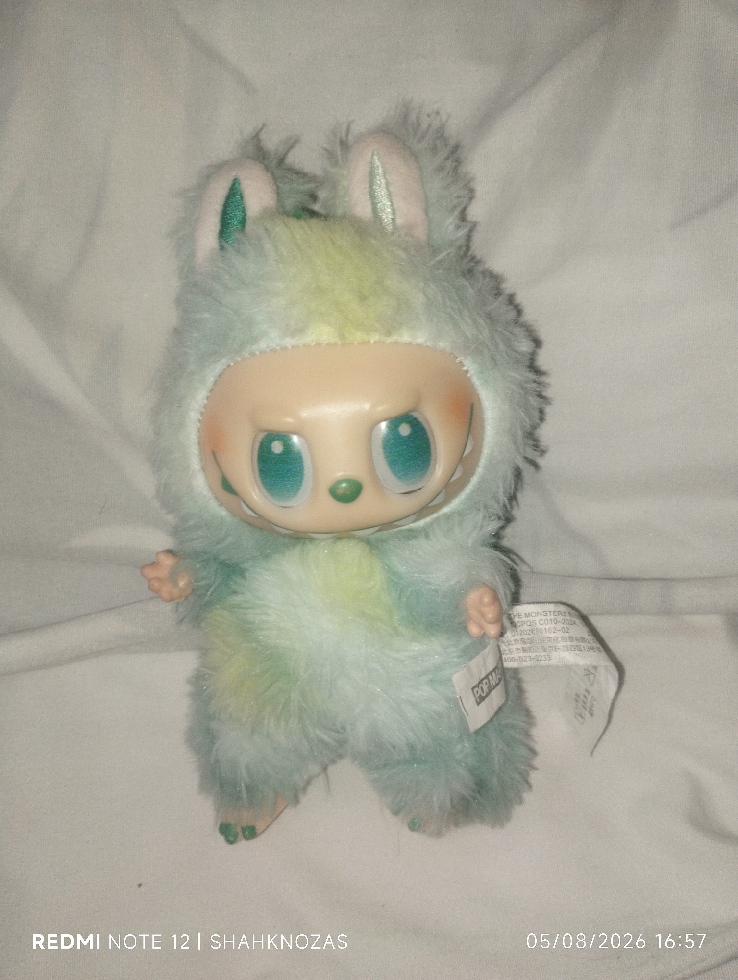 A stuffed toy, approximately 10 inches tall, is positioned on a white, wrinkled fabric surface. The toy has a pale plastic face with large, teal eyes, a small green nose, and a wide mouth revealing small white teeth. It is covered in soft, fluffy fur that transitions from a pale blue at the bottom to a yellow-green on the chest and head, with pink and teal accents on its rabbit-like ears. The toy is wearing a light blue furry hood. A white tag with black text is attached to its right side. The overall impression is of a whimsical, possibly alien-like creature. The timestamp "05/08/2026 16:57" and "REDMI NOTE 12 | SHAHKNOZAS" are visible in the lower right and left corners respectively, indicating the image was taken on August 5, 2026, at 4:57 PM. The location provided, Chibintepa, Uzbekistan, is not discernible from the visual information in the image.