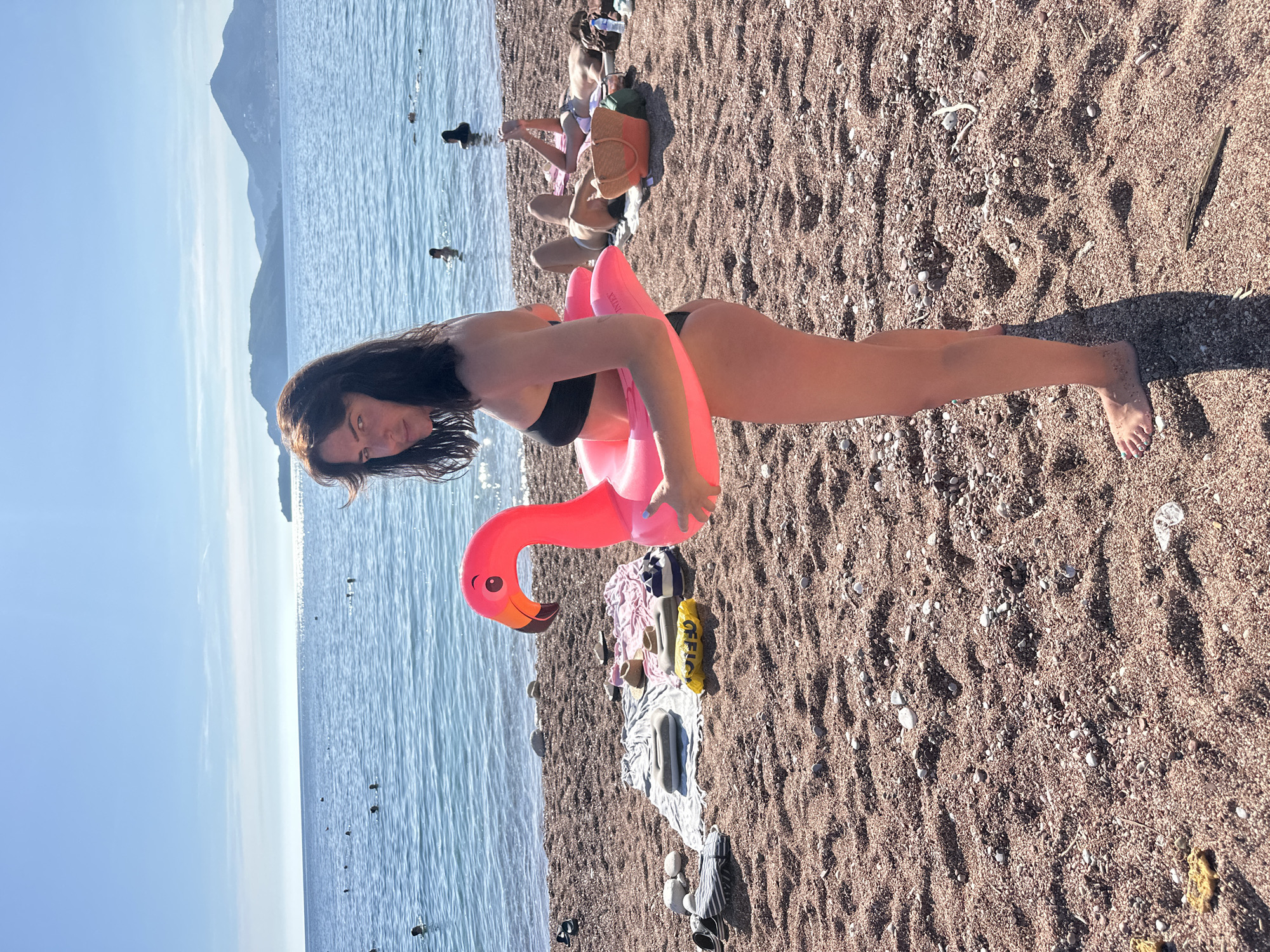 A young woman with dark hair is standing on a pebble beach in Sveti Stefan, Montenegro, holding a bright pink inflatable flamingo. She is wearing a black bikini and looking over her shoulder at the camera with a neutral expression. The beach is populated with other beachgoers, some swimming in the clear blue water, others lounging on towels and beach chairs. The sea stretches out to the horizon, meeting a clear blue sky dotted with a few wispy clouds. In the distance, mountains are visible. There are various beach items scattered around the sand, including towels, hats, a bag, and flip-flops. The sun is shining brightly, casting shadows on the sand.