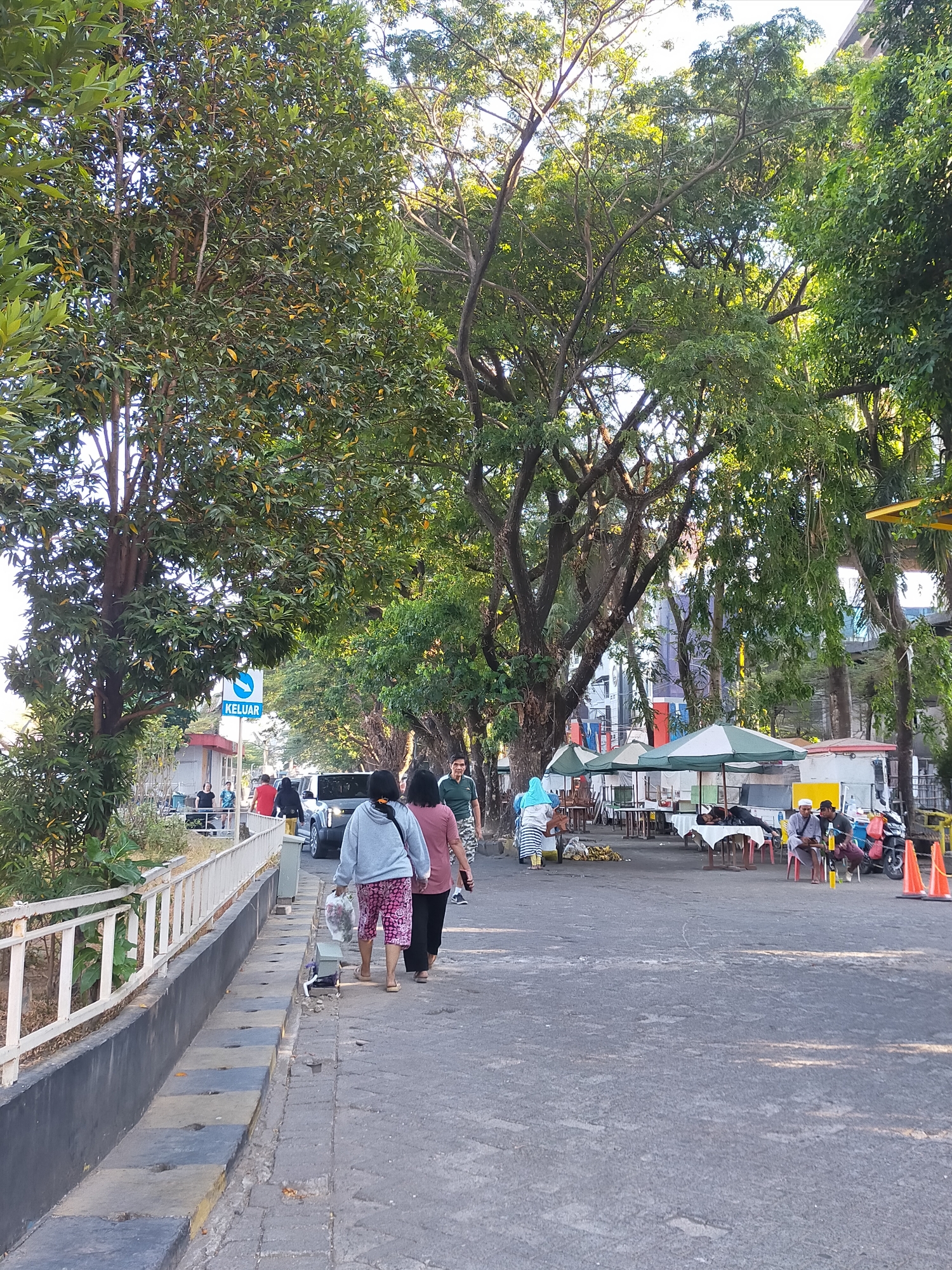 This image captures a street scene in Makassar, Indonesia, with a focus on the sidewalk and a portion of the road. The foreground features a gray concrete sidewalk with a white railing along its edge, bordering a raised planter area with some greenery. Two women are walking away from the camera on the sidewalk, one in a gray hoodie and patterned shorts, the other in a maroon shirt and dark pants. Several other individuals are visible further down the sidewalk and on the road.
The road itself is paved and appears to be a two-lane street. Several vehicles are parked or moving on the road, including a dark SUV. To the right of the road, there are several outdoor stalls or vendors set up with tables and umbrellas, suggesting a marketplace or food court area. Some people are seated at the tables.
The background is dominated by large trees with dense green foliage, casting dappled shadows on the ground. A building, possibly a multi-story structure, is visible behind the trees. The overall lighting suggests it is late afternoon or early evening, with warm sunlight filtering through the leaves. A blue and white sign with the word "KELUAR" (Indonesian for "Exit") is visible on a pole near the sidewalk. The environment appears to be a busy urban setting with elements of both commerce and natural greenery.