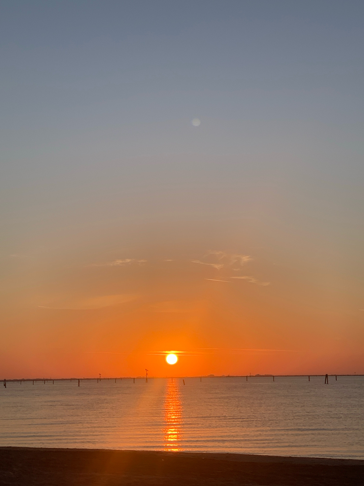 The image depicts a serene sunset over the waters of Comacchio, Italy. The sun, a brilliant orb of orange, is low on the horizon, casting a shimmering golden path across the calm sea. The sky transitions from a soft blue at the top to vibrant shades of orange and pink as it nears the horizon. Numerous vertical posts, likely markers or part of a historical fishing system, are visible in the water, spaced evenly across the scene. In the distance, a faint line of buildings suggests the presence of the city. The foreground shows a dark, sandy shore. A small, pale circular object, possibly the moon or a distant planet, is visible in the upper portion of the sky. The overall atmosphere is one of tranquility and natural beauty.