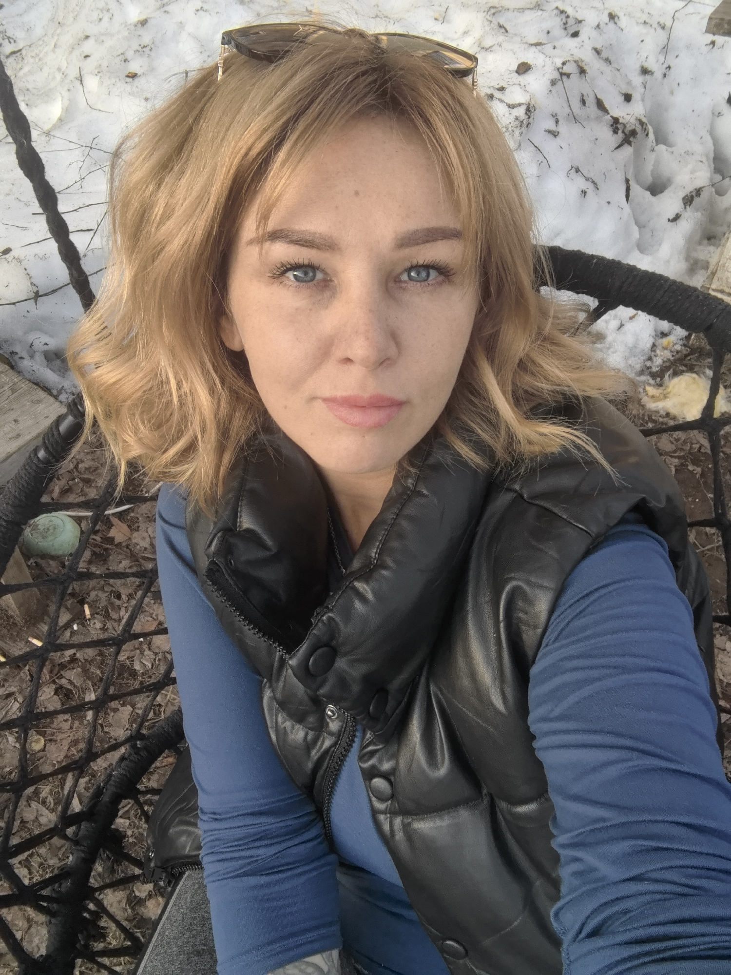 A blonde woman with fair skin and freckles, wearing sunglasses on her head, a black puffer vest over a blue long-sleeved shirt, is seated in a black woven chair. The chair sits on a patch of ground covered with dry leaves, snow, and bits of trash, including what appear to be cigarette butts and a green saucer. In the background, there is a snow-covered area with dark branches visible, and a piece of weathered wood. The woman appears to be taking a selfie. The location is plausibly in Noviy, Russia, although nothing directly in the image confirms the exact location.