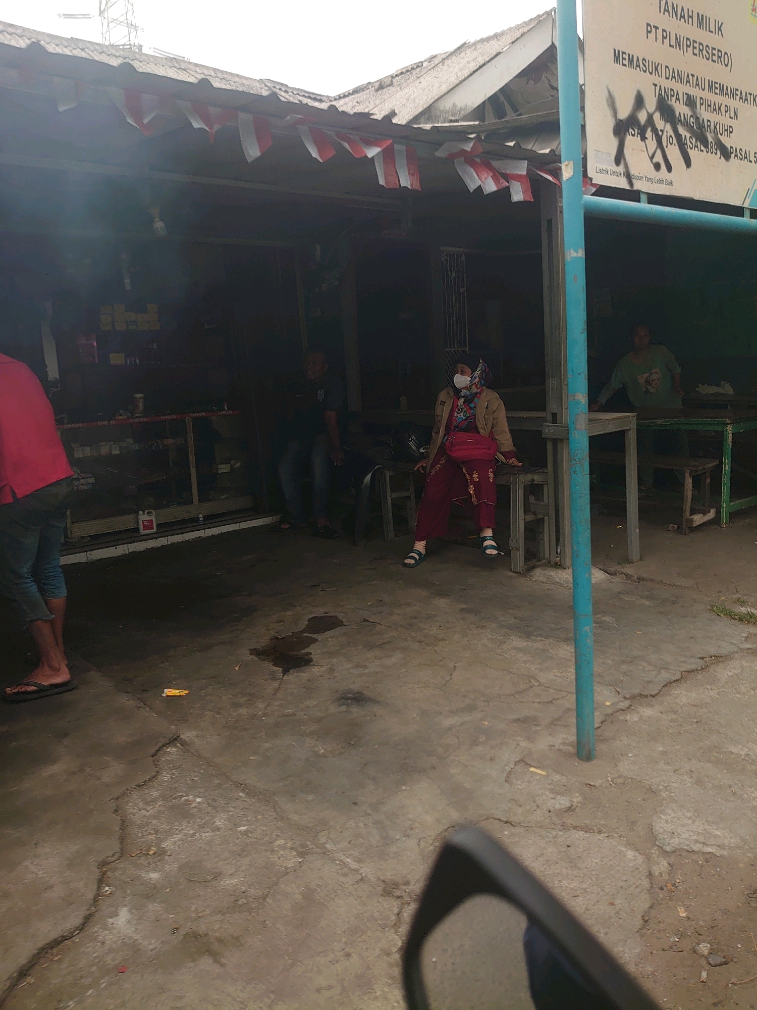 The image depicts an outdoor scene in what appears to be Depok, Indonesia, characterized by a dilapidated storefront and its surrounding area. In the foreground, a person's legs and feet are visible, wearing flip-flops, with the lower part of a vehicle's interior reflected in the bottom right corner, suggesting the photo was taken from inside a car.
The storefront itself is covered by a corrugated metal roof, adorned with red and white Indonesian flags strung along the edge. The structure appears aged and somewhat neglected. Inside the dimly lit storefront, shelves are stocked with various items, though they are indistinct.
In the mid-ground, several individuals are present. A woman wearing a light brown jacket, a patterned headscarf, and a red mask is seated on a bench, leaning slightly forward. She is dressed in a maroon outfit and has a red crossbody bag. To her left, a person wearing a dark shirt and blue jeans is seated. Further to the right, another person in a light green shirt stands behind a table.
The ground outside the storefront is paved with concrete, showing signs of wear, cracks, and some water or oil stains. A bright blue pole stands prominently in the mid-ground. In the background, a large sign is visible, partly obscured by graffiti, with visible text mentioning "TANAH MILIK PT PLN (PERSERO)" and other legal disclaimers. The overall atmosphere is casual and appears to be a local commercial establishment.