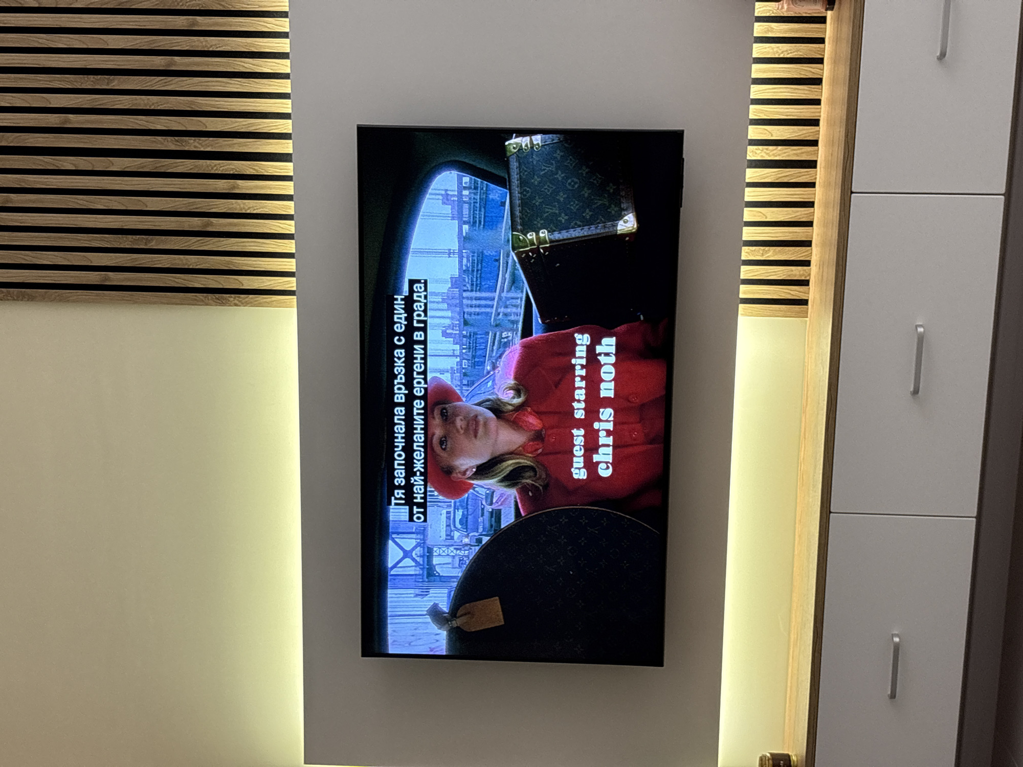 The image shows a television screen mounted on a wall in what appears to be an interior setting. The TV displays a scene with a woman in a red coat and hat. Text on the screen, presumably subtitles or captions, are in Bulgarian and English, with “guest starring chris noth” visible. A Louis Vuitton bag is also displayed on the screen. To the left of the TV is a wall decorated with horizontal wooden slats. To the right, there is a light colored cabinet with silver handles. The wall around the TV is illuminated with soft lighting. This is likely in Sofia, Bulgaria due to the use of Bulgarian text on the television.