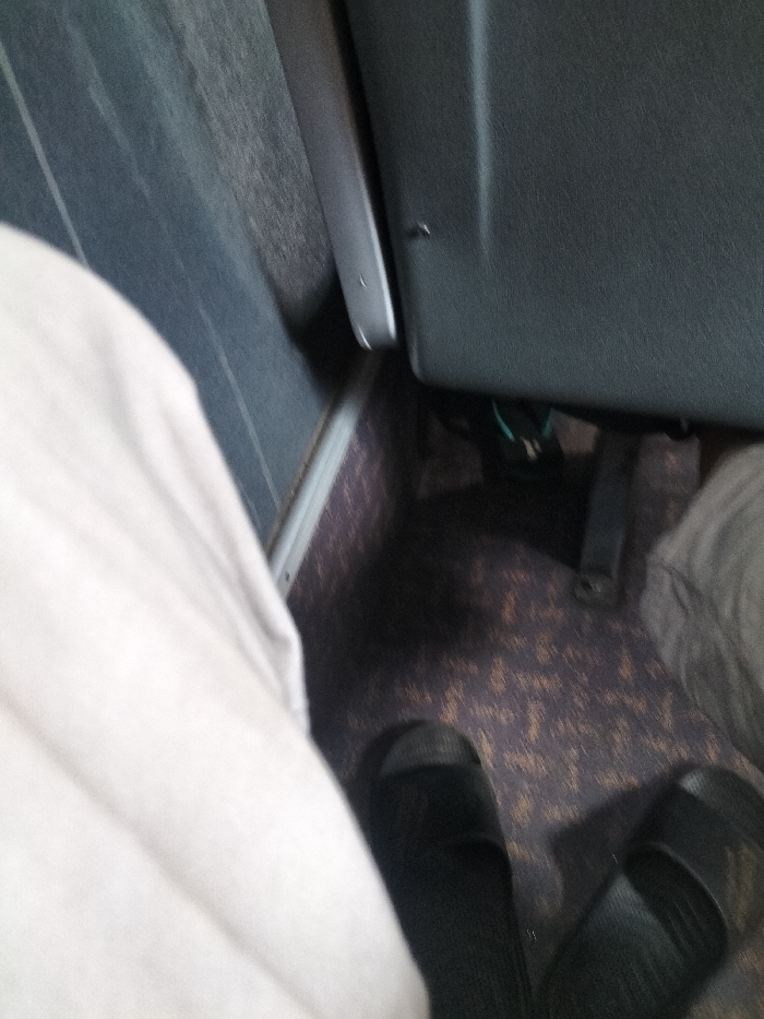 The image is taken from the perspective of a person sitting in a bus. The person's right leg is visible, with the foot clad in black slippers. The leg is resting on the aisle, and the person's right hand is also visible holding the bus seat. The bus seat is covered in dark gray fabric. The floor of the bus is carpeted in brown with a pattern of repeating shapes. To the right of the aisle, another person's legs and part of the seat are visible. The scene is located in the city of Kebomas, Indonesia, at the coordinates -7.168 latitude and 112.6 longitude.