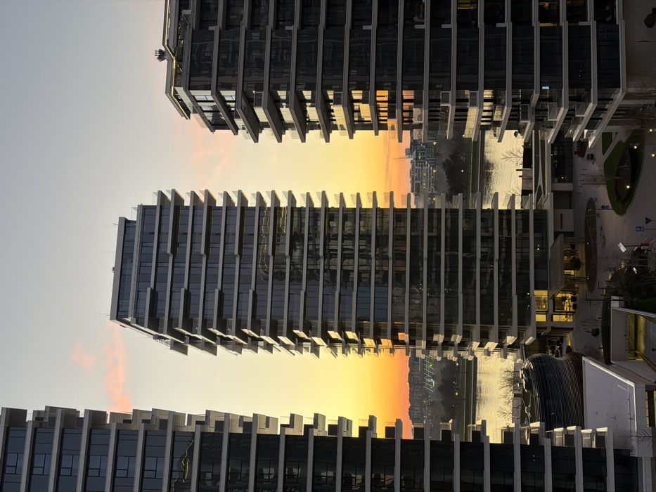 The image displays a modern urban landscape in the Belgrade Waterfront district of Belgrade, Serbia, captured during sunset or twilight. Three contemporary high-rise buildings are the primary subjects. The central building, a tall structure with a dark glass facade and numerous balconies featuring glass railings, is flanked by partially visible similar buildings on the left and right. The Sava River flows in the midground, reflecting the sky's gradient which transitions from a vibrant orange and yellow near the horizon to a pale blue overhead, with subtle pink clouds.
At ground level, to the left of the central building and adjacent to the river, a distinctive curvilinear multi-level structure with a blue and brown striped facade is visible. Below the buildings, a landscaped pedestrian area is evident, comprising paved pathways, small, bare deciduous trees, and an entrance to an underground parking garage. Distant urban architecture is discernible on the far bank of the Sava River. Artificial interior lights are illuminated in various windows of the buildings and at ground-level entrances. No individuals are visible within the scene.