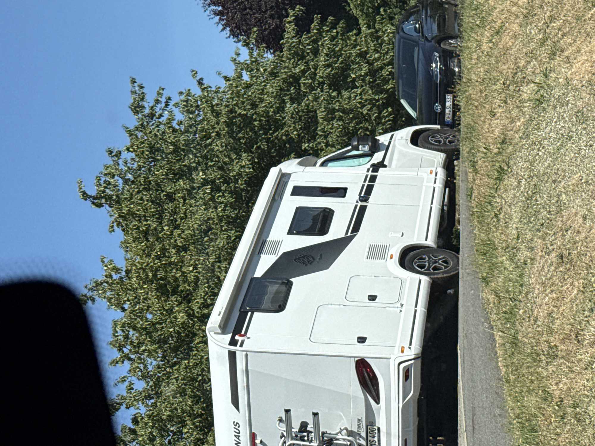 The image shows a white recreational vehicle (RV) parked on the side of a road in Bad Sachsa, Germany. A black Volkswagen Polo is parked next to the RV. The RV has a bicycle rack mounted on the back. The license plate of the Volkswagen Polo is visible and reads "OHA SL 333". Trees and a bright blue sky are in the background. The scene appears to be a sunny day.