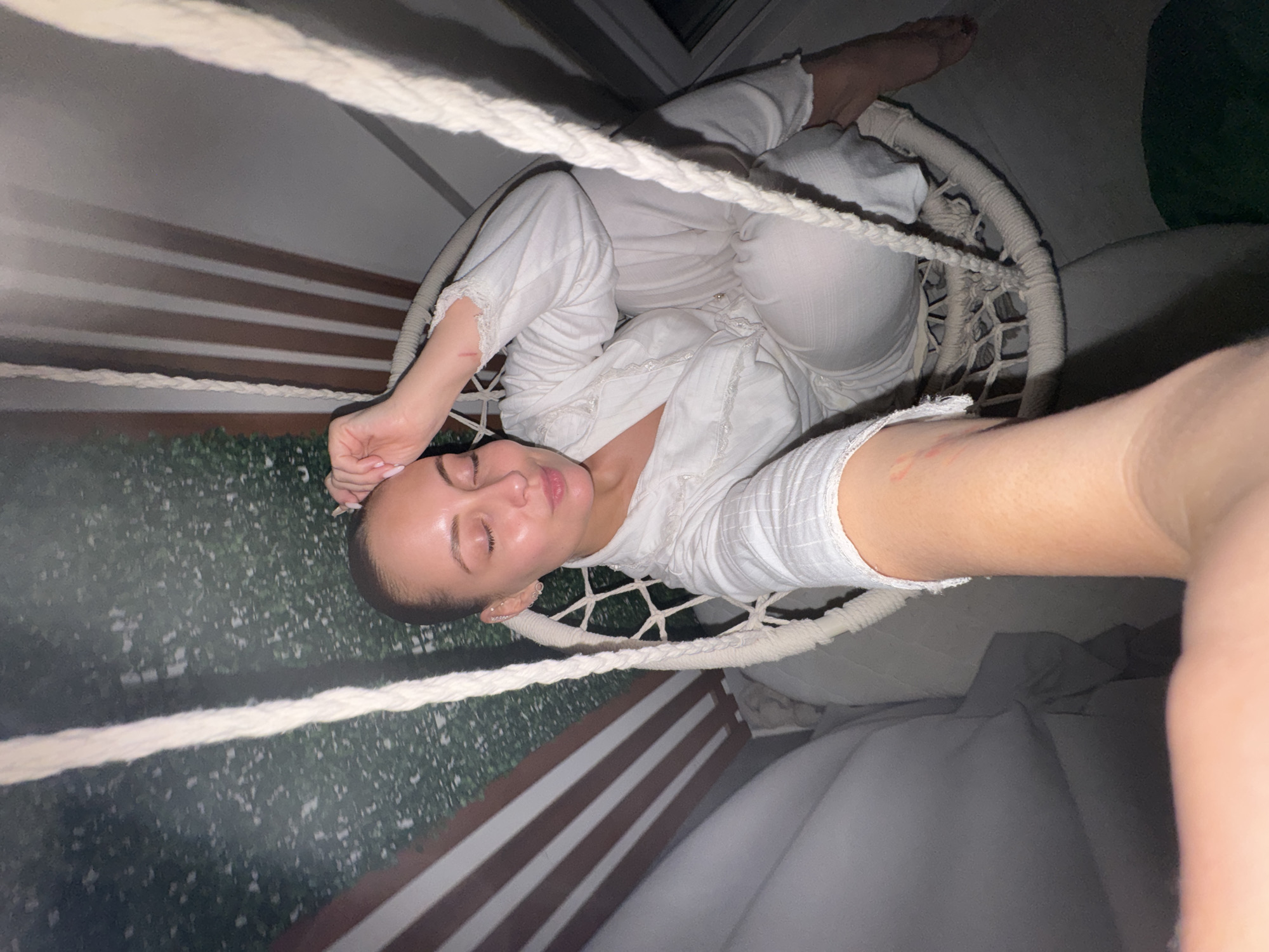 An individual, likely female, is captured in a low-angle shot while resting in a white, woven hanging chair. She is dressed in a relaxed, matching white long-sleeved top and pants. Her eyes are closed, and her head is tilted, conveying a sense of peace or relaxation. Her hair is pulled back, and her fingernails have light pink polish. A small reddish mark or scratch is visible on her right forearm.
Thick white ropes, which suspend the chair, are prominent in the foreground and background. The setting appears to be indoors or on a sheltered balcony in Chaikovsky, Russia, indicated by the dim lighting, suggesting evening or night. In the background, there's a wall featuring horizontal dark and light brown slats, possibly wood paneling or a decorative railing. Below this, a textured green surface resembling artificial foliage or a hedge wall is visible. A window pane can be seen in the upper left portion of the image. No visible text is present.