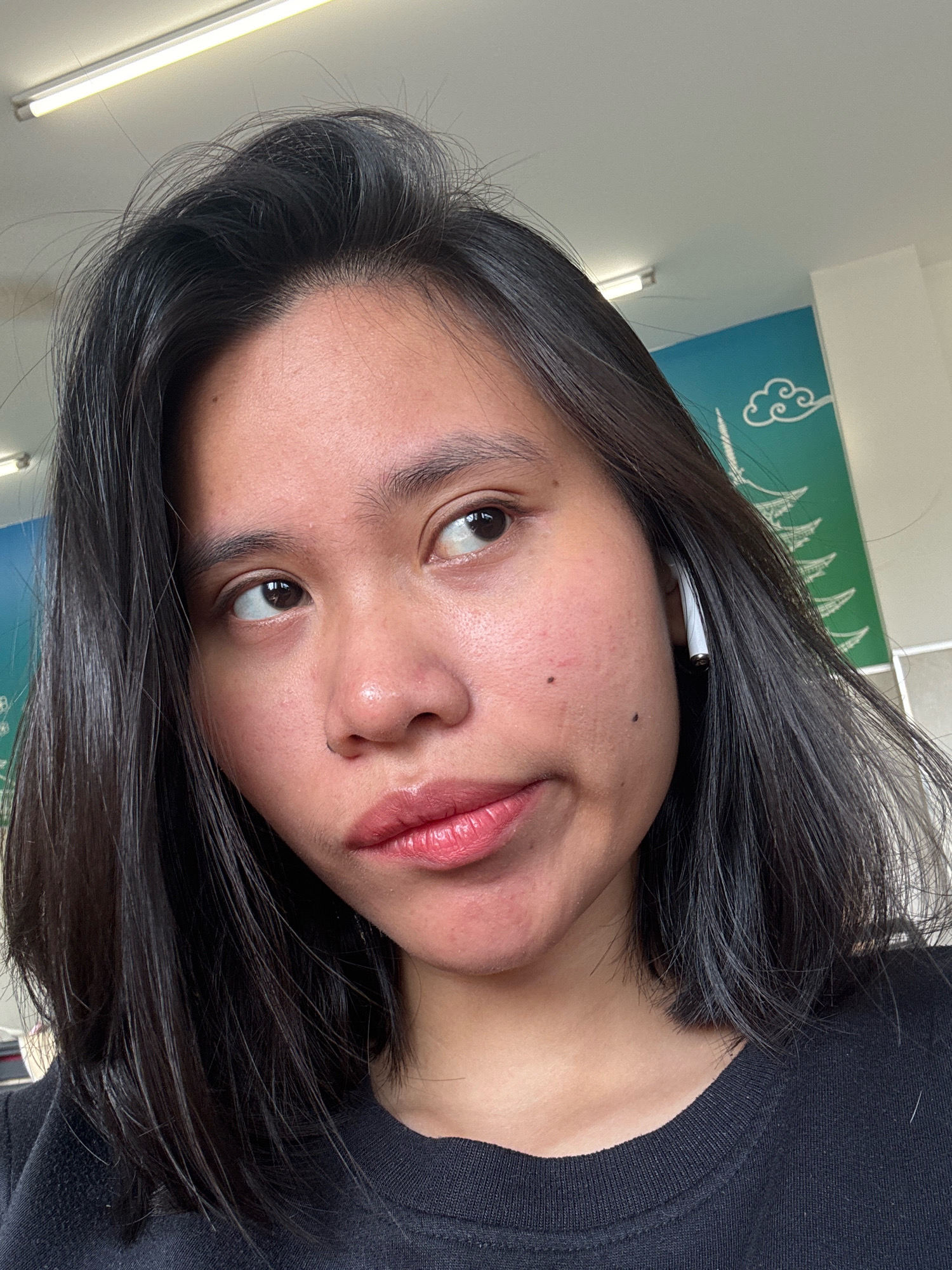 A medium close-up shot features a young East Asian woman with dark hair, wearing a black shirt. She has a white earbud in her right ear. The background shows a white ceiling with fluorescent lighting, and a portion of a teal-colored wall with a white cloud and tree graphic. The image appears to be taken indoors, possibly in an office or classroom setting in East Jakarta, Indonesia.