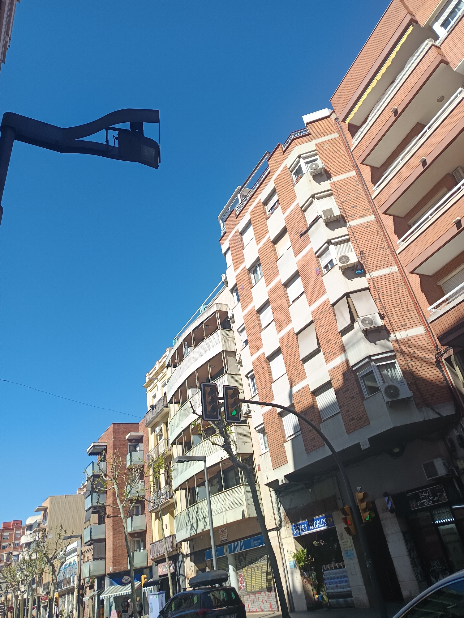 The image captures a street scene in L'Hospitalet de Llobregat, Spain. A variety of multi-story residential buildings line the street, showcasing a mix of architectural styles and materials, including brick, white facades, and balconies. Air conditioning units are visible on the exteriors of several buildings.
Traffic lights are present, with one showing a green light. Streetlights and bare trees are also visible. The lower levels of the buildings house commercial establishments, with shop signs and awnings. A barber shop is on the corner.
A black car is parked along the street. The sky is clear and blue, suggesting a sunny day.
