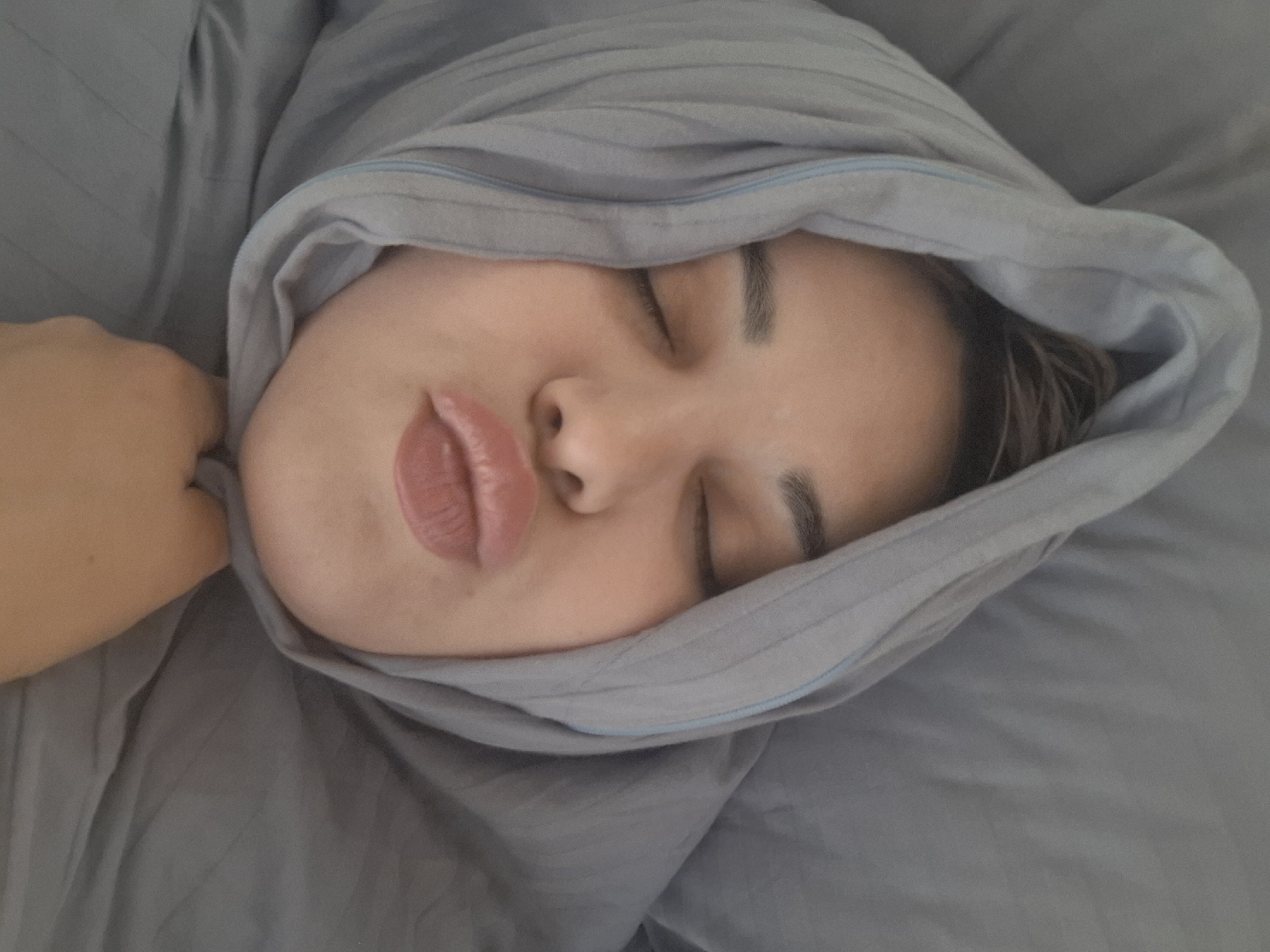 A person is lying in bed, covered in a grey blanket with a light blue trim. The person's eyes are closed, and their lips are puckered. A hand is visible in the lower left of the frame. The location is unknown.
