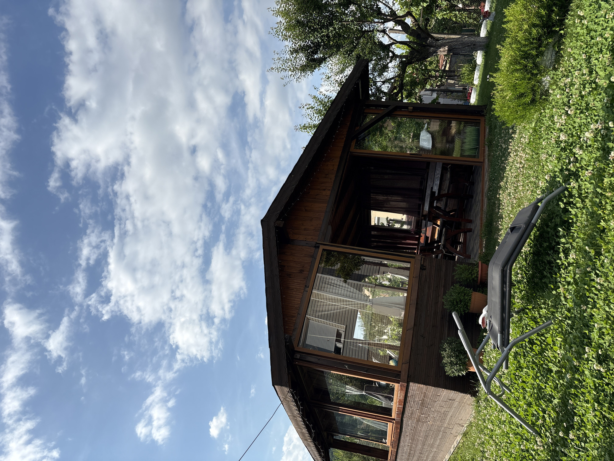 This image captures an outdoor scene in Polena, Bulgaria, featuring a multi-sided wooden structure, likely a gazebo or sunroom, situated on a verdant lawn. The structure is constructed from dark brown stained wood, including horizontal paneling on its lower section, and is topped with a dark shingled roof. Large glass windows and a glass door comprise the visible facade, revealing internal light-colored blinds or curtains, potted plants, and interior furniture consisting of a table and chairs. Small decorative string lights are visible along the roof eaves on the left side of the structure.
The surrounding ground is a dense green lawn intermixed with numerous small white clover flowers. In the immediate left foreground, a grey, metallic-framed foldable lounge chair with a dark fabric sling rests on its side. Several potted green bushes are arranged directly in front of the wooden structure. To the right of the building, a large, mature deciduous tree with green foliage stands, partially obscuring the structure's far right side. A long, manicured green shrub is visible in the middle ground on the right.
The sky occupies the upper portion of the frame, displaying a clear blue with scattered white cumulus and cirrus clouds. A thin black cable extends diagonally across the upper left quadrant of the sky. The scene is illuminated by natural sunlight, casting visible shadows. No individuals or animals are present in the image.