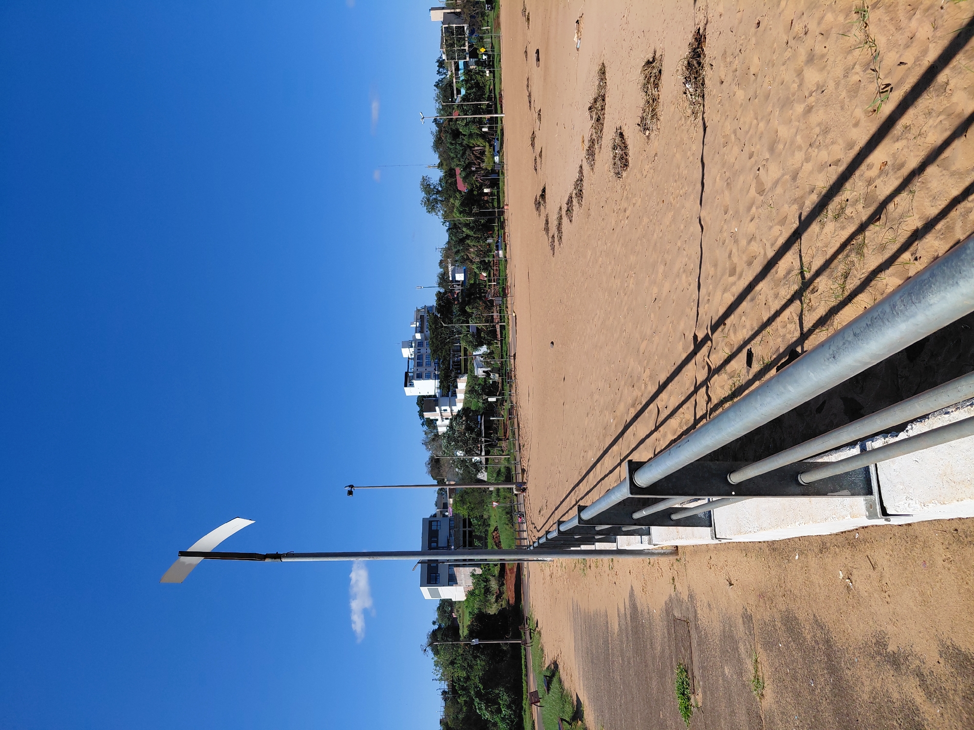 The image captures a scene at the Costanera de Encarnación, Paraguay, on a clear day. In the foreground, a metal railing runs along the edge of a sandy area. To the left of the railing, a concrete pathway is visible, with some sparse green vegetation growing in cracks. The sandy area, which appears to be a beach or park, has several small piles of debris scattered across it, likely consisting of dried leaves and twigs. In the middle ground, a lamppost stands tall, casting a shadow on the sand. Further back, a row of trees lines the area, with modern buildings visible behind them, indicative of the urban environment of Encarnación. The sky is a vibrant, cloudless blue, suggesting good weather.