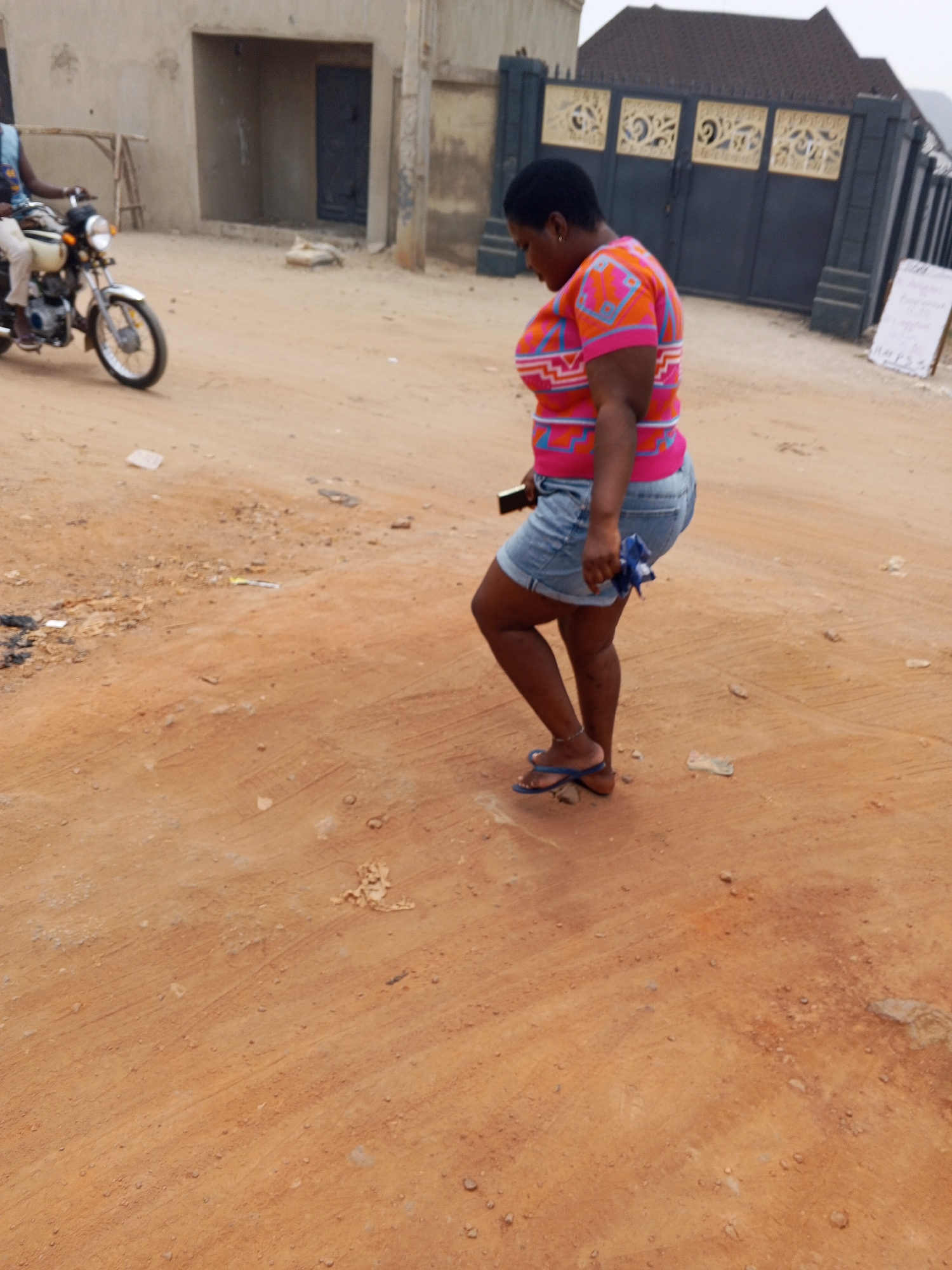 The image depicts a woman walking on a dirt road in Adonkolo, Nigeria. She is wearing a pink and orange patterned top, denim shorts, and blue flip-flops. She is holding a smartphone in one hand and a small blue object in the other. The road is uneven and dusty. In the background, there is a motorcycle with a rider, a building with a doorway, and a dark metal gate with decorative elements. A white sign stands near the gate.