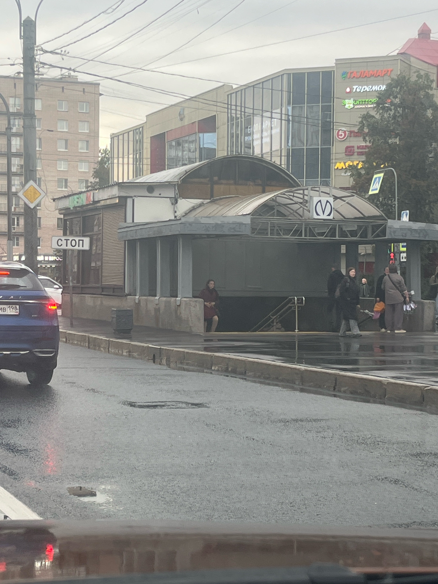 This is a photograph taken on a cloudy day in Saint Petersburg, Russia, near a metro station entrance. In the foreground, a blue car is stopped on a wet asphalt road. The road is reflecting the overcast sky and the brake lights of the car. To the right of the car, a sidewalk and curb are visible, also wet from rain.
Behind the sidewalk is the entrance to a metro station, characterized by a covered walkway with a distinctive "M" logo for the metro. People are standing under the cover of the station entrance, some appearing to be waiting or walking. One person in a dark coat is walking towards the entrance, and another person in a red coat is seated near the entrance.
Further in the background, a multi-story building with modern glass architecture is visible, featuring prominent signage for "TAJAMART", "Teremok", and other businesses. A traditional brick residential building is also present to the left of the modern structure. A traffic signal with a "STOP" sign is also mounted on a pole near the metro entrance. The overall environment suggests an urban setting in Saint Petersburg, likely during a time of overcast weather and recent rainfall.