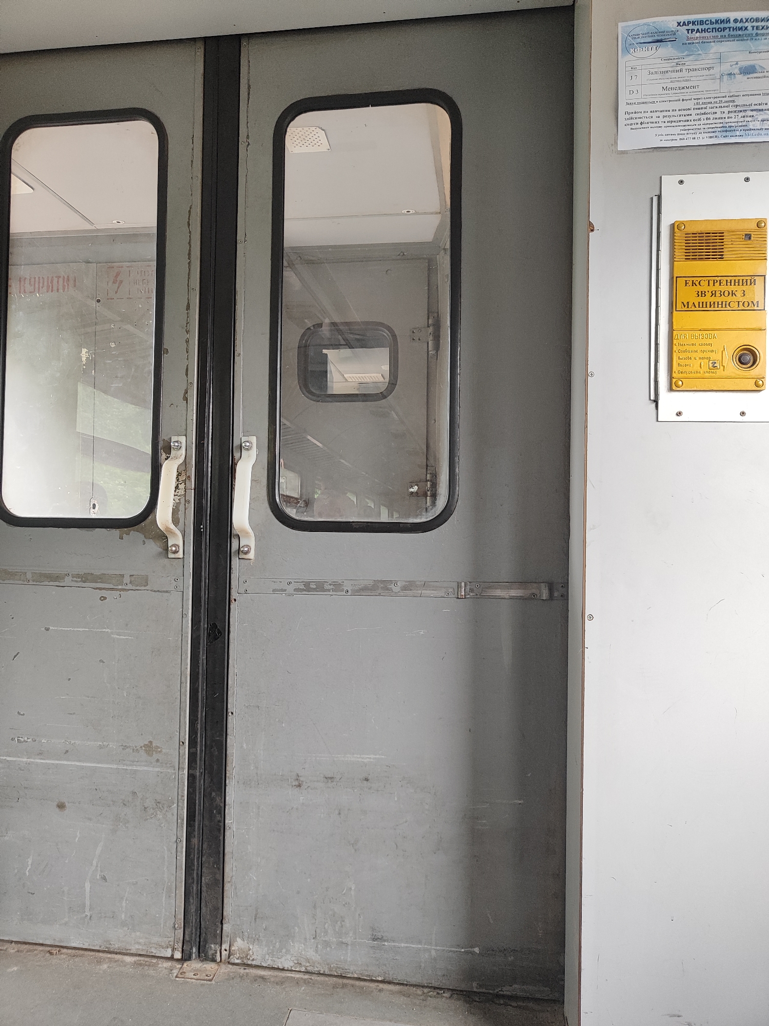 The image shows a close-up of two gray, weathered, double doors with rectangular windows. The doors have white handles. To the right of the doors, there is a white panel with a yellow emergency call button and intercom system. The panel has text in Cyrillic, indicating it is an "EMERGENCY CALL TO THE DRIVER" system. There is also a paper document attached to the wall above the yellow panel, with Cyrillic text and a seal. The document appears to be an official certificate or permit related to transportation. The floor in front of the doors is a concrete surface. The overall scene suggests an entrance to a public transportation vehicle or facility, possibly a train or subway car, given the emergency call system and the official documentation. The lighting is diffused, indicating an indoor or overcast outdoor setting.
