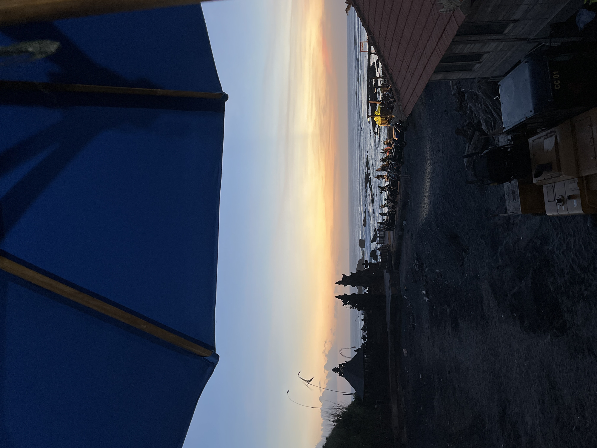 The image captures a serene coastal scene in Mengwi, Indonesia, during what appears to be a sunrise or sunset. A large blue umbrella occupies the left foreground. The sky transitions from pale blue to a warm, orange hue, reflecting on the sea. The coastline is lined with trees and a structure resembling a temple or traditional building. The right side of the image shows a building with a red tile roof and dark, possibly volcanic, sandy ground. There are also vehicles and some structures alongside the shoreline, suggesting a populated area.
