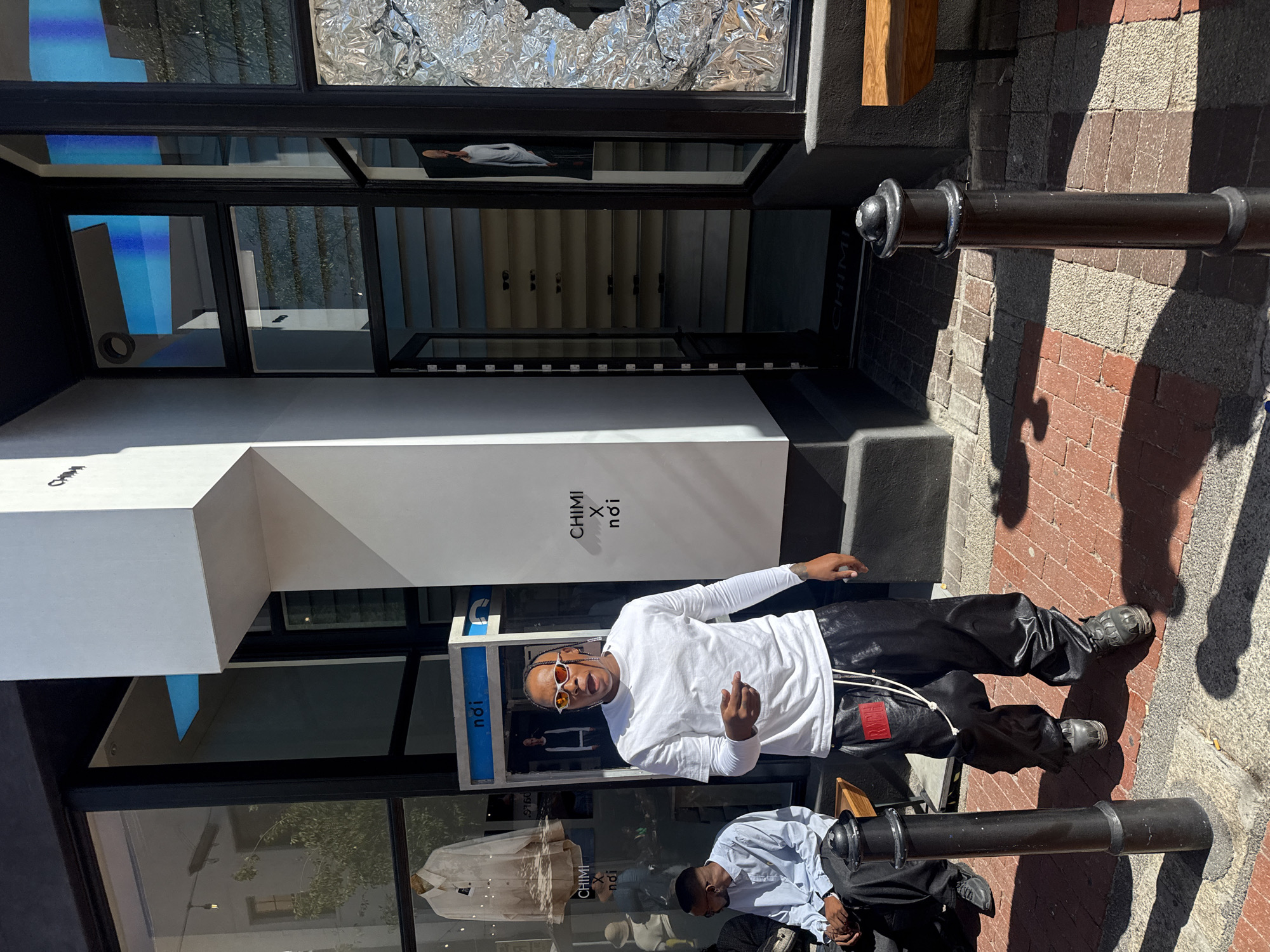 The image displays an urban street scene on Bree Street, Cape Town, South Africa, during daylight hours. A prominent modern retail storefront occupies the background, featuring large black-framed glass windows and an angular white architectural projection bearing the black text "CHIMI x nōi." The left display window showcases a light-colored long-sleeved garment on a hanger, while the right window contains silver crinkled foil material and a visible poster depicting a person in a white top. An open glass door serves as the store entrance, revealing interior shelving consistent with eyewear display.
In the foreground, a dark-skinned male subject, estimated mid-20s to early 30s, walks towards the viewer and slightly left. He has braided hair and wears orange-red mirrored sunglasses with white frames, a white long-sleeved t-shirt, and shiny black baggy pants adorned with a red rectangular patch and white drawstring. Dark, robust footwear is visible. His mouth is open, and his right arm is slightly raised and extended forward in a distinct gesture. A tattoo is visible on his left forearm.
To the left of the primary subject, a second dark-skinned male is seated on a wooden bench or ledge. He wears a light blue collared shirt and dark trousers, with small dark-rimmed glasses, and appears to be looking left. A black metal bollard stands between the two individuals, slightly right of the frame's center. The ground is paved with reddish-brown and light-brown bricks, interspersed with grey concrete, casting strong, defined shadows indicative of direct sunlight.