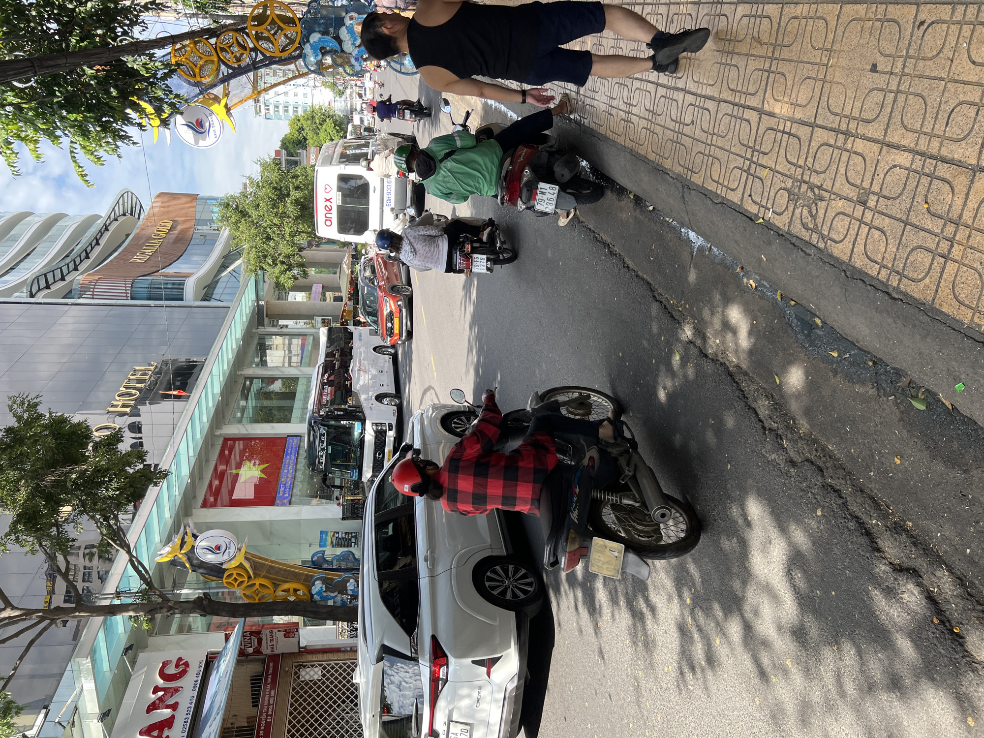 The image depicts a street scene in Nha Trang, Vietnam, on a sunny day. In the foreground, a person on a red and black plaid shirt is riding a motorcycle, their face partially obscured by a red helmet and a mask. To the left, a white SUV is parked, and further down the street, traffic is visible, including several other motorcycles, a red car, a white van with "anex" branding, and two buses. One bus displays "LUXURY PALACE." The buildings lining the street are modern, featuring glass facades and signage for businesses like "HOTEL" and "REGALIA GOLD." Decorative elements, including yellow metallic structures with circular motifs and blue wave-like designs, are visible along the sidewalk. The sidewalk itself is paved with patterned tiles, and a man in a black tank top and blue shorts is walking away from the camera on it. The road surface shows signs of wear, with cracks and patches. Trees with green foliage are interspersed throughout the scene, casting shadows on the road and sidewalk. The sky is partly cloudy with blue patches.