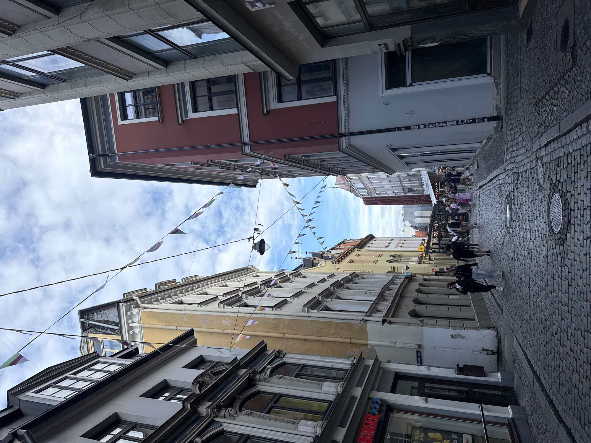 The image shows a street scene in Riga, Latvia, with historic buildings lining a cobblestone street. In the foreground, two people, a male and a female, are walking down the street away from the camera. They are both wearing casual clothing. The street is lined with buildings that appear to be from the 19th or early 20th century, with intricate architectural details. Some of the buildings have shops or restaurants on the ground floor. The sky is partly cloudy, and the overall lighting suggests it is daytime. There are strings of triangular flags strung across the street between some of the buildings, possibly indicating a festival or event. In the background, more people are visible walking and gathered outside establishments. The street itself is made of gray cobblestones, with some manhole covers visible. The image captures a lively, European city atmosphere.