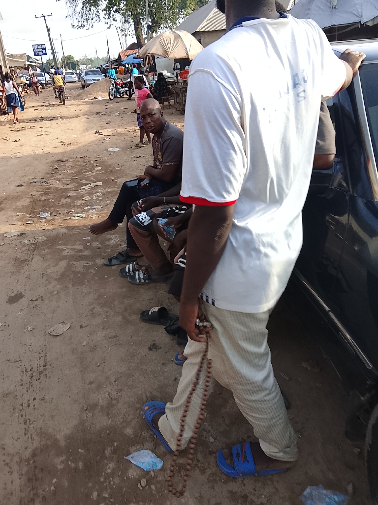 The image depicts a street scene in an unspecified area of Abuja, Nigeria. The environment consists of a dusty, unpaved road with litter scattered around. Several people are visible, some sitting and others walking or riding motorcycles. A group of men is seated along the side of a black car; one man stands, leaning on the car. There are commercial activities with shops and stalls lining the streets. A sign reading "RUFAIDAN YOGHURT & FURA" is seen in the background. People wear casual clothing, and sandals are the predominant footwear.