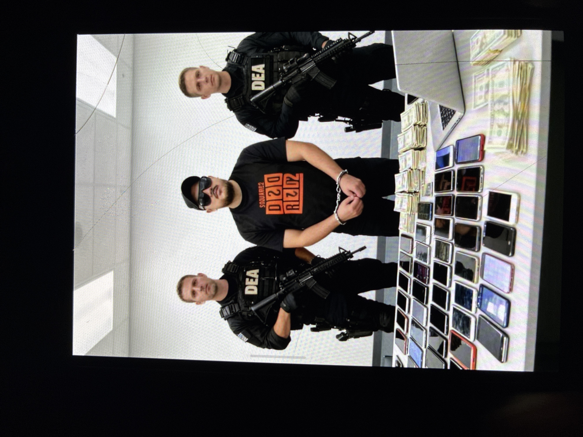 The image depicts an indoor scene with three individuals: two law enforcement officers identified by "DEA" patches on their vests, armed with rifles, and a man in handcuffs. The man in handcuffs is wearing a black T-shirt with orange text, sunglasses, and a black cap. A table holds stacks of U.S. currency, a laptop, and numerous cell phones. The background is a light-colored wall with ceiling tiles. No specific location in Kalitinovo, Bulgaria, is evident in the image.