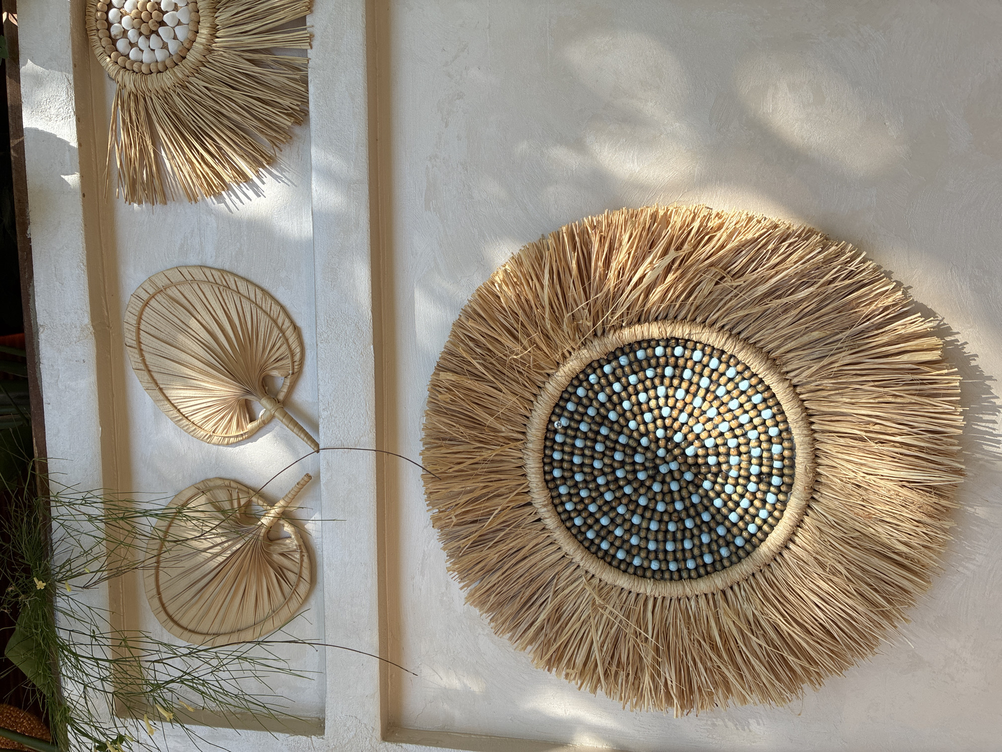 The image displays a wall with decorative items made of natural materials. The wall itself is textured and painted a light, neutral color. There are three circular or fan-shaped decorations attached to the wall.
The top left features two fan-shaped decorations, appearing to be made of dried palm leaves, fanned out and attached to a handle-like base. They are positioned side-by-side.
To the top right, there is a circular decoration with a fringe of straw-like material radiating outwards. The center of this decoration is composed of smaller, round elements, possibly beads or shells, in shades of white and light brown.
Dominating the lower portion of the image is a larger, circular wall hanging. It also features a fringe of natural, straw-like material. The central area of this large decoration is intricately arranged with small beads. These beads are primarily a light brown or wooden color, interspersed with a vibrant, light blue bead, creating a patterned effect.
Green, wispy foliage hangs into the frame from the top left, casting shadows on the wall and decorations. Sunlight streams in from the right, creating distinct areas of light and shadow, highlighting the textures of the wall and the decorative elements.
The style of decoration suggests an outdoor or semi-outdoor setting, possibly a patio, balcony, or a restaurant/cafe interior with a natural, bohemian aesthetic. The presence of greenery and the use of natural materials are characteristic of such environments. This specific scene is located on a wall in Alanya, Turkey, likely part of an establishment that embraces a natural, earthy, or bohemian design theme. There are no people or animals visible in the image.