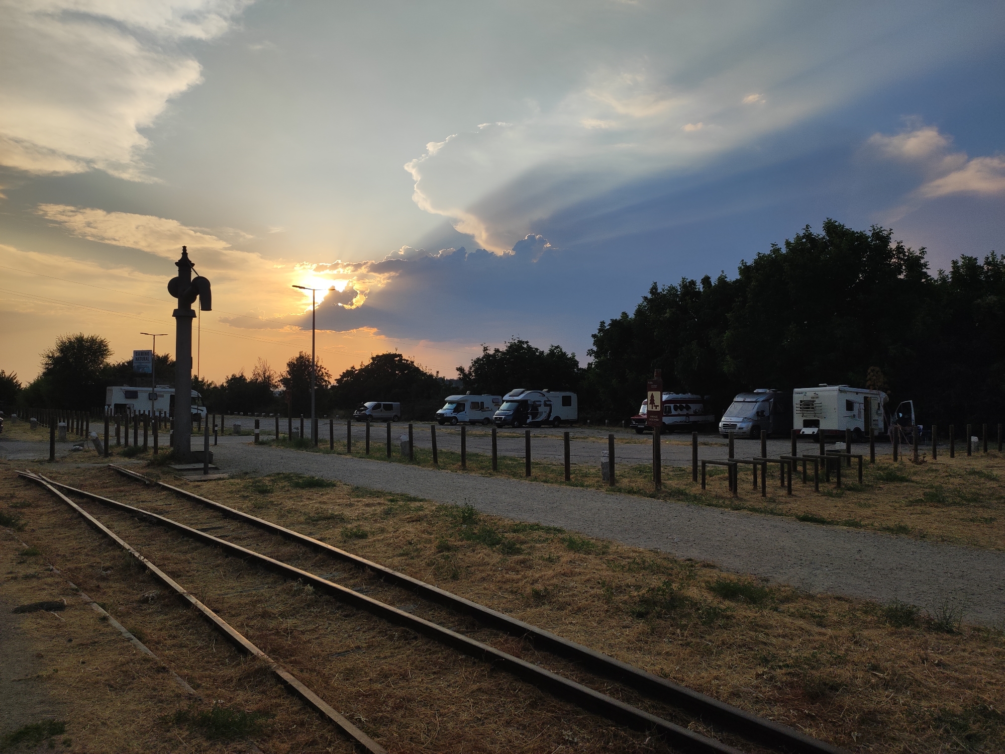 This image depicts a scenic view at dusk in Béjar, Spain. The foreground features a set of disused railway tracks, with dry, golden grass growing around them. Beyond the tracks, a gravel parking area is visible, lined with wooden posts. Several campervans and RVs are parked in this area, suggesting it might be a campsite or an area designated for recreational vehicles.
In the background, a dense line of dark green trees forms a natural barrier, with some of the trees silhouetted against the sky. The sky itself is a dramatic canvas of changing light. The sun is low on the horizon, peeking through and behind large, wispy clouds, casting a warm, orange glow and creating visible sunbeams. The clouds range from bright white near the sun to darker blues and grays further away, indicating a dynamic and potentially changing weather pattern.
A notable object in the mid-ground is an old-fashioned railway signal, a dark, metallic structure standing tall next to a lamppost. There's also a street lamp on the right side of the image. A small sign with text is partially visible on a post near the trees, but the text is illegible. The overall mood is serene and tranquil, with the warm light of the setting sun creating a peaceful atmosphere. The presence of the old railway infrastructure combined with modern campervans hints at a location that bridges history and contemporary leisure.