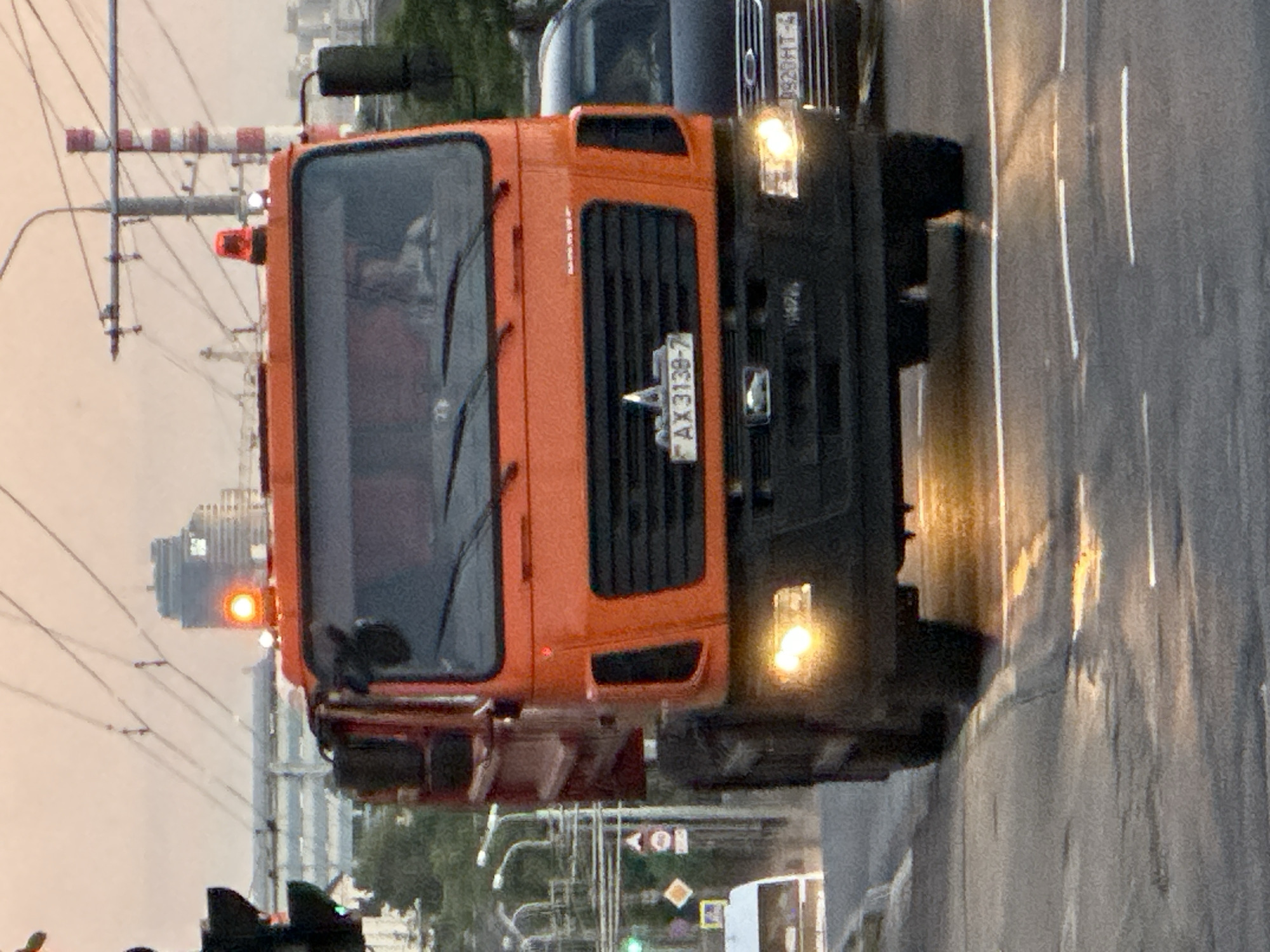 A horizontally-oriented, medium-shot captures a bright orange MAZ utility truck, likely a road maintenance vehicle, driving on an urban street. The truck, facing the viewer, features a dark grey grille, front bumper, and fenders. The license plate, visible on the grille, reads "AX 3139-7," indicating Belarusian registration. Above the truck, a second vehicle, partially visible, is identified by a Belarusian license plate reading "9920HT-4." The road surface is wet, reflecting light from the vehicles and the streetlights below.
In the background, an overhead railway structure with red and white barriers is visible. Further back, a distant cityscape appears, with a prominent building featuring a dark, reflective facade. The overall lighting suggests either dusk or dawn, with a soft, muted color palette. The setting is identified as Minsk, Belarus, based on the license plate.
