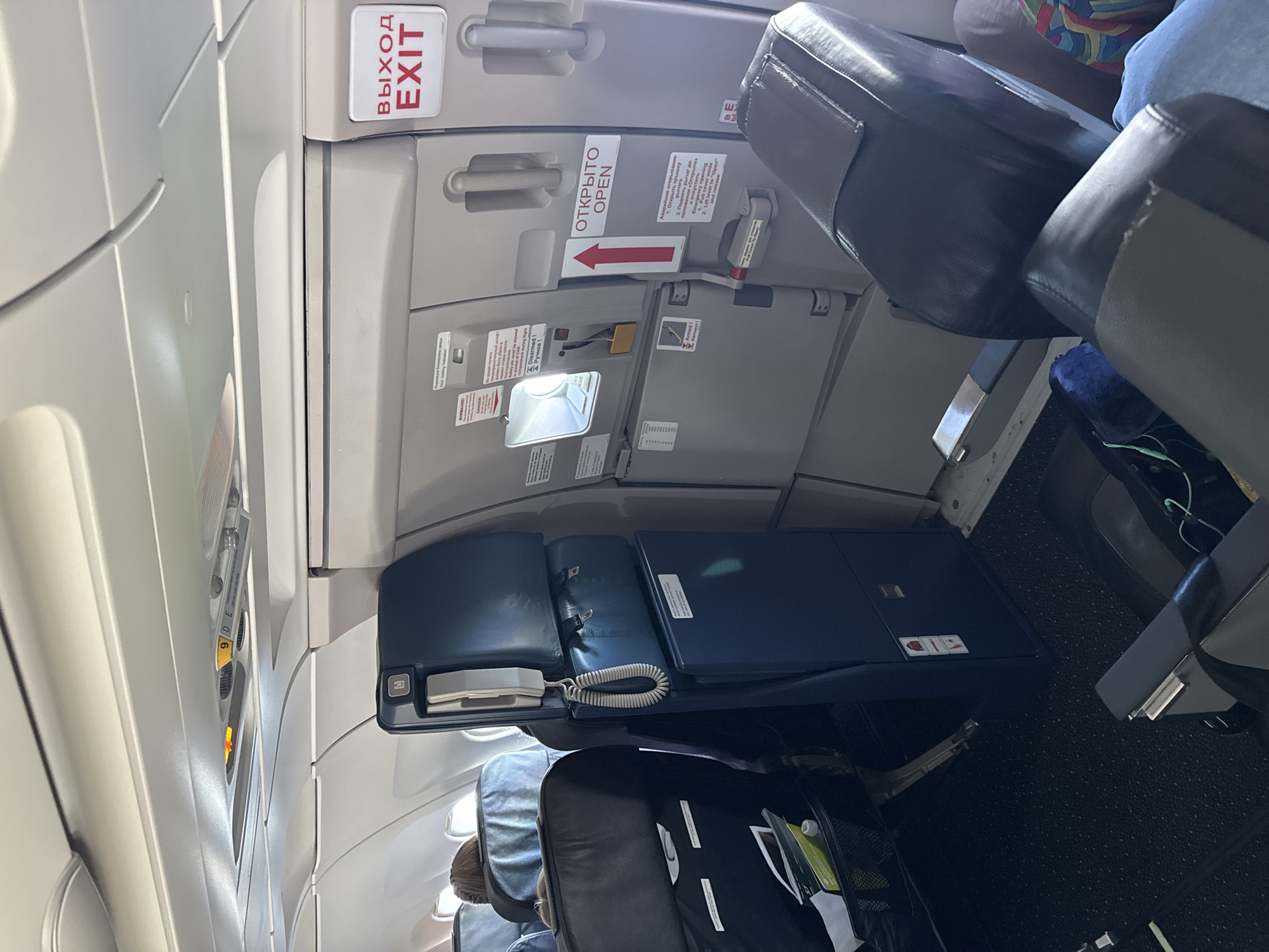 The image shows the interior of an airplane cabin, specifically a view of seats and an aircraft door. The scene is captured from a low angle, looking upwards and towards the front of the aircraft. In the foreground, dark-colored airplane seats with grey armrests are visible, with one seat occupied by a person wearing blue jeans and a colorful top. The seats have seatbelts.
The middle ground reveals a section of the aircraft's interior wall, which includes an exit door. This door is marked with bilingual signage in Russian and English. A white sign with red text reads "ВЫХОД EXIT," indicating an emergency exit. Below this, on the door itself, is a red arrow pointing upwards with the Russian text "ОТКРЫТО" and the English word "OPEN" below it. There are also various labels and instructions on the door, some appearing to be safety warnings and operational procedures. A white telephone handset is attached to a blue panel next to the door.
Further back, rows of airplane seats are visible, some with headrests. Above the seats, the overhead compartments and lighting fixtures of the cabin are visible. On the ceiling, a yellow warning light is illuminated. The floor of the cabin is covered in dark, textured carpeting.
The image is likely taken in the city of Ob, Russia, given the Cyrillic text on the signage, which suggests Russian airline operations. However, the image itself does not provide explicit confirmation of the specific location within Ob, other than it is on an airplane. The overall environment is sterile and functional, characteristic of an aircraft cabin.